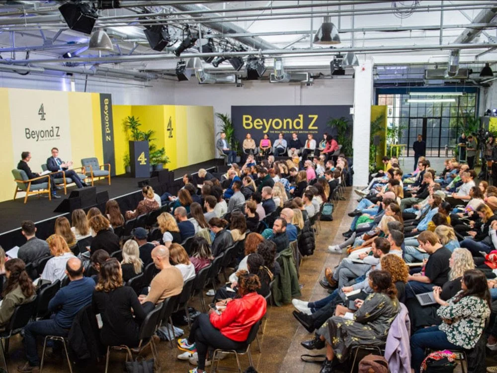 A large conference or panel discussion event with many attendees seated and some on stage, with a backdrop reading 'Beyond Z', in an industrial-style venue with plants and large windows.