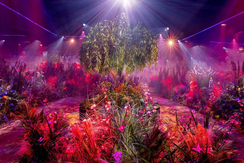 Colorful indoor garden with a central tree, vibrant flowers, and lush greenery illuminated by stage lights and spotlights.