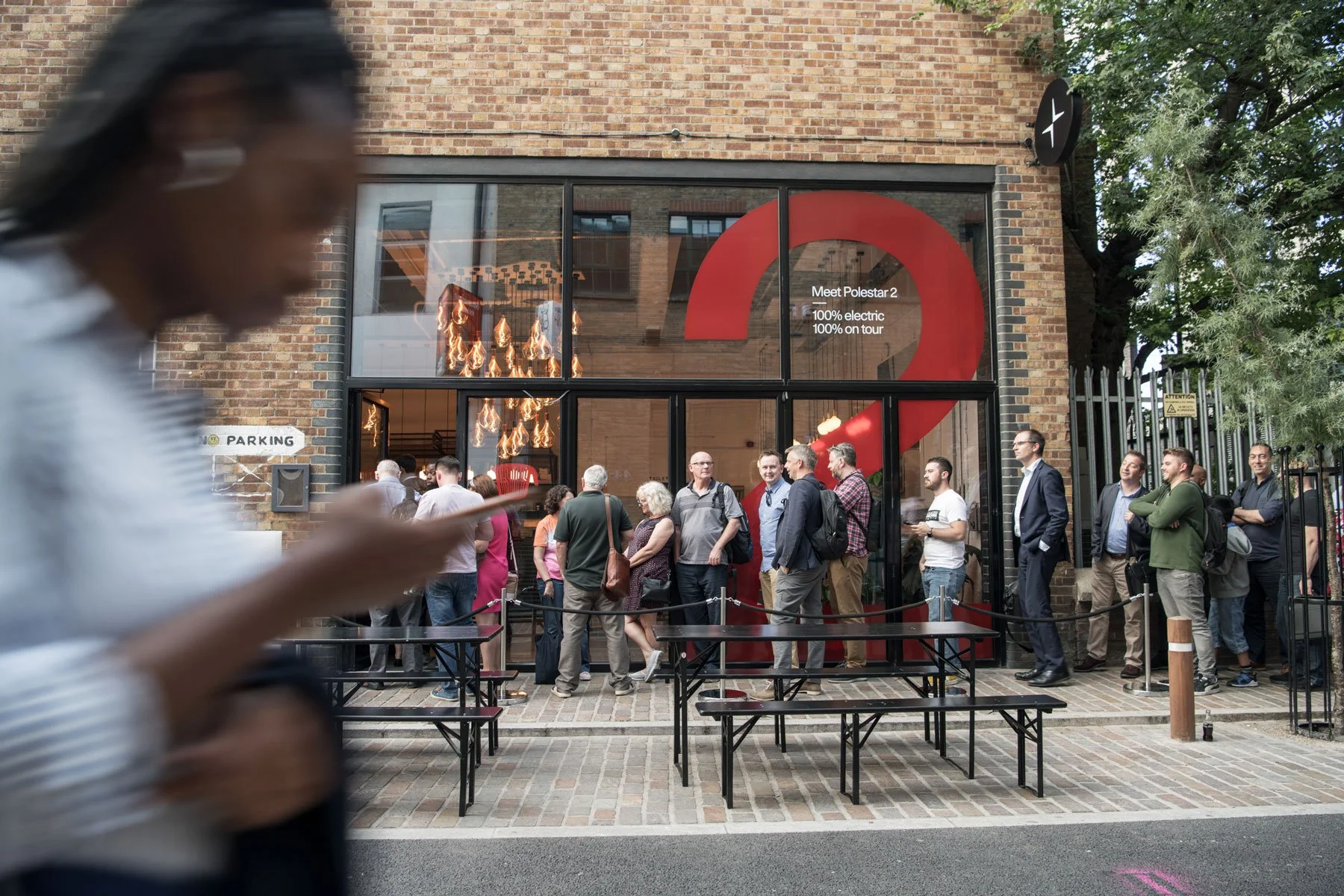 A line of people waiting outside a modern building with a large red question mark and text indicating they are attending an event for Polestar 2, a 100% electric car. The building has glass windows showing interior lighting, and there are trees and a