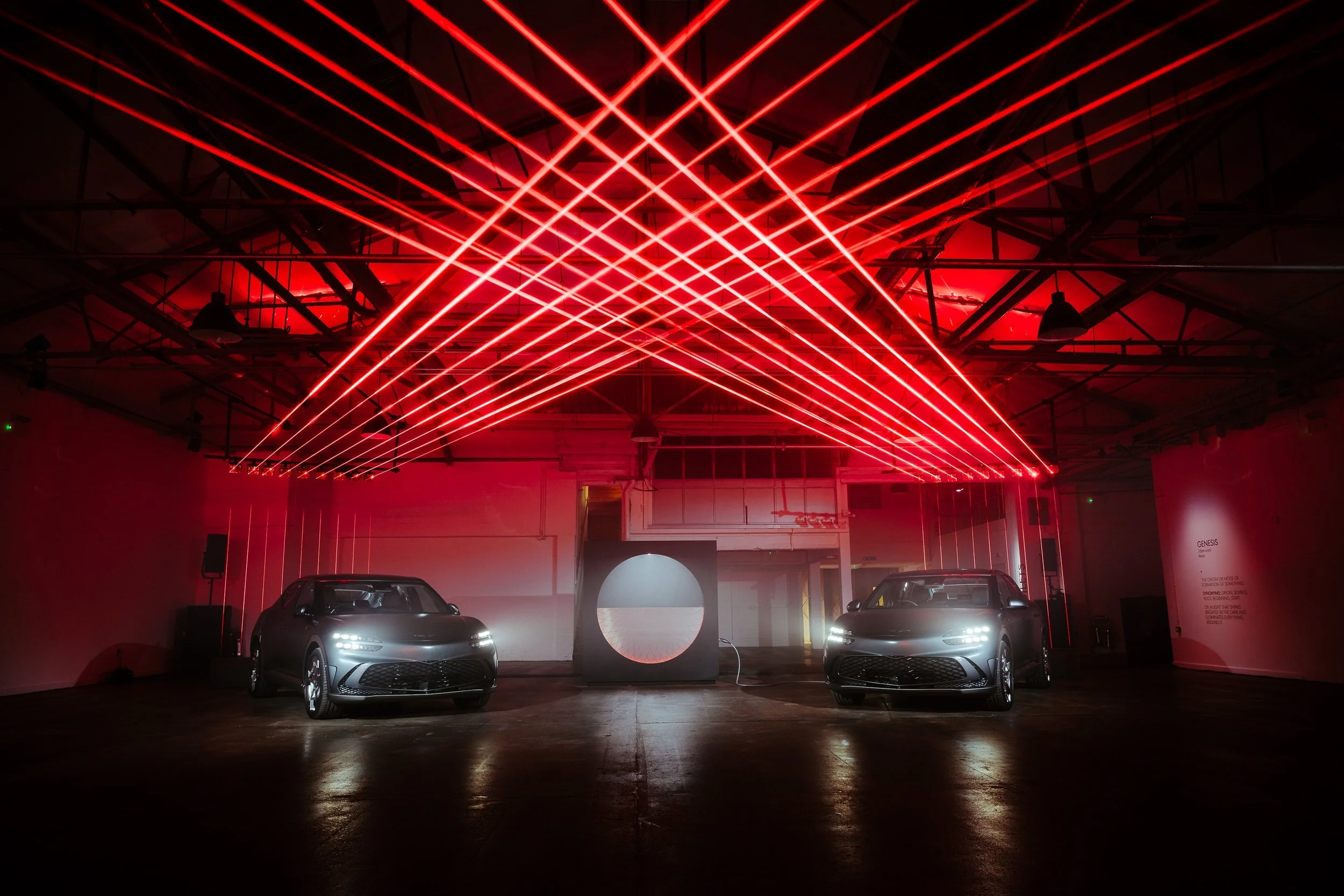 Two silver cars parked in a dark indoor space with red laser lights forming an abstract pattern above them.