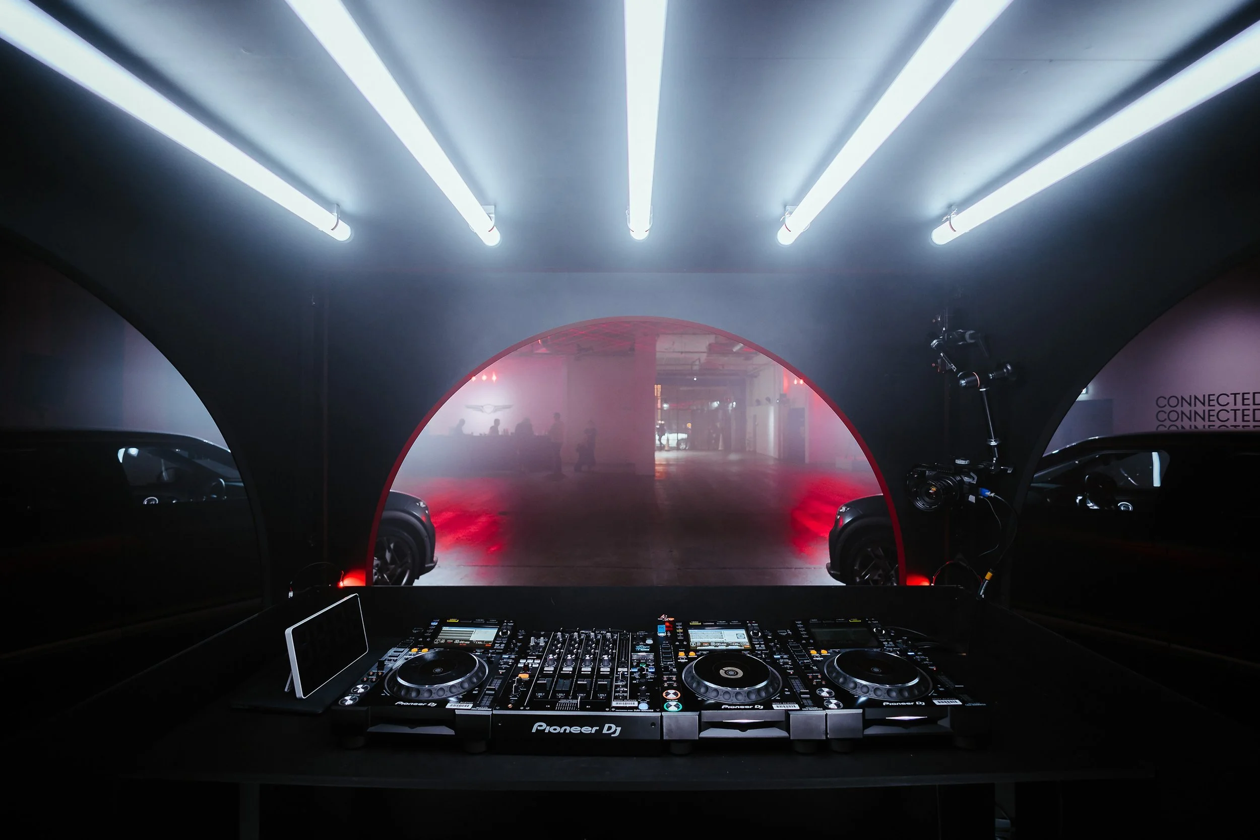 A DJ setup with turntables, mixer, and a tablet on a table in front of a foggy, church-like archway with neon lights on the ceiling, and cars parked in the background.