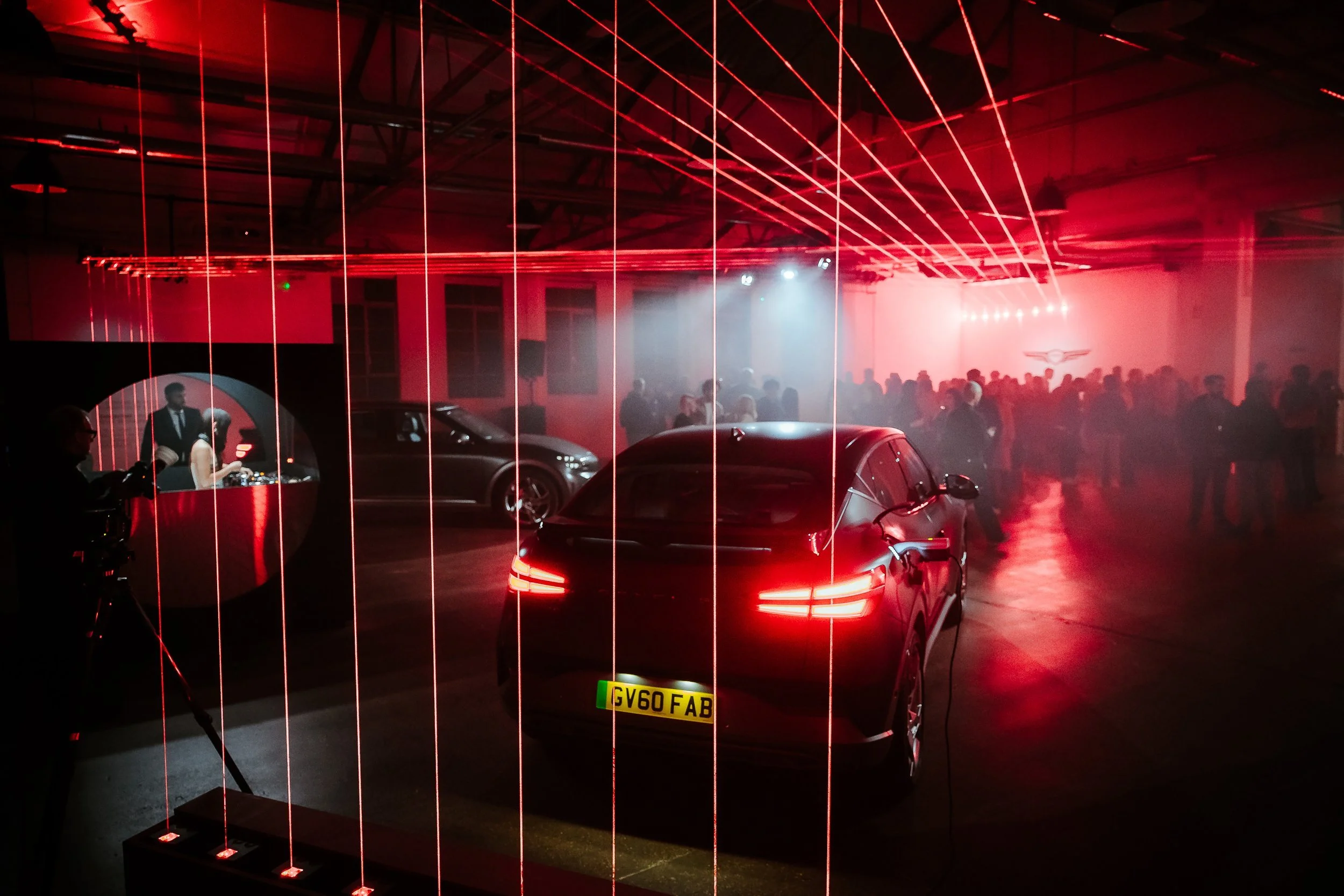 An indoor car exhibition with red lighting, featuring a black luxury car in the foreground, a DJ booth on the left, and a crowd of people in the background.