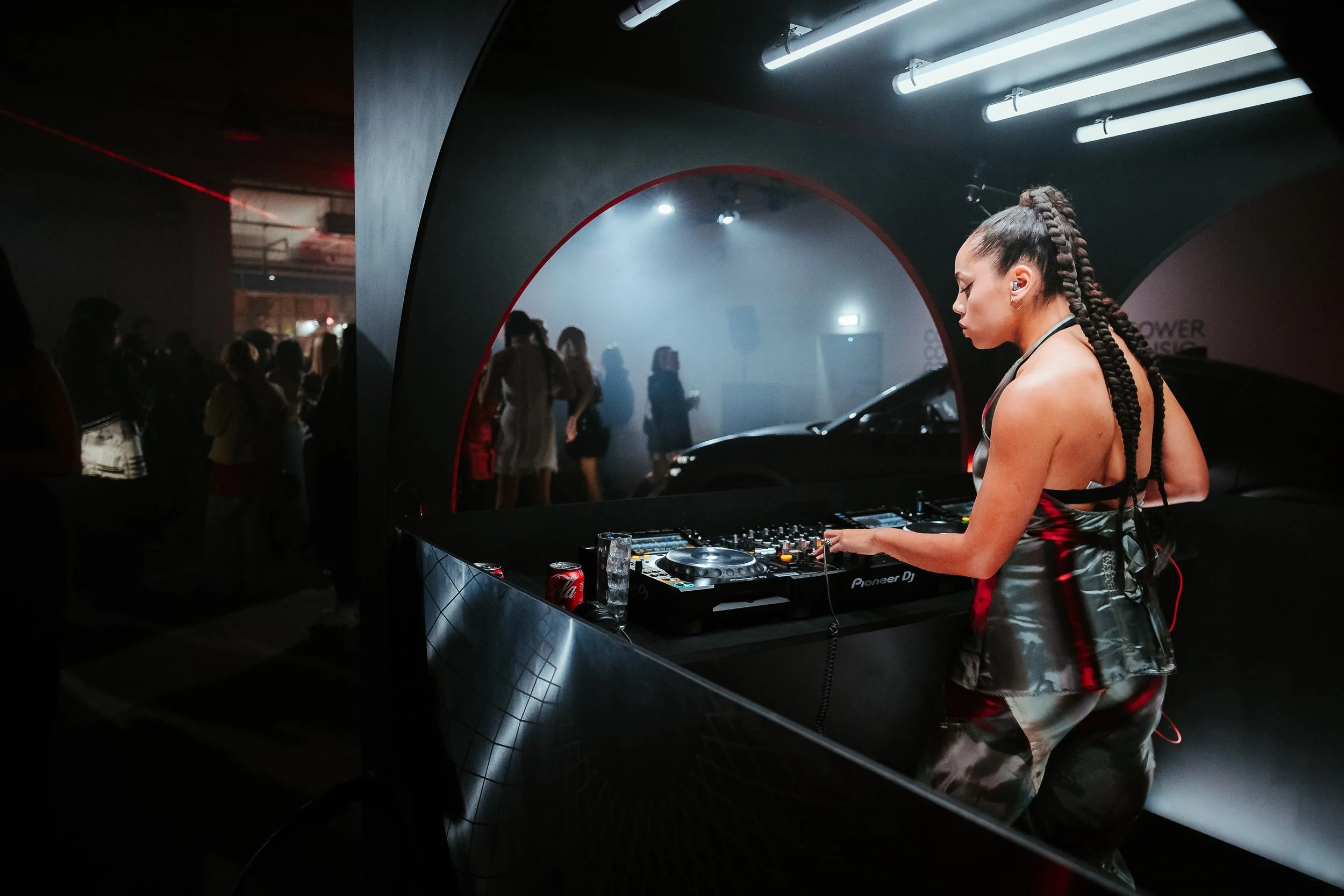 A female DJ with braided hair wearing a shiny black outfit performing at a nightclub or concert, with a crowd of people dancing in the background.
