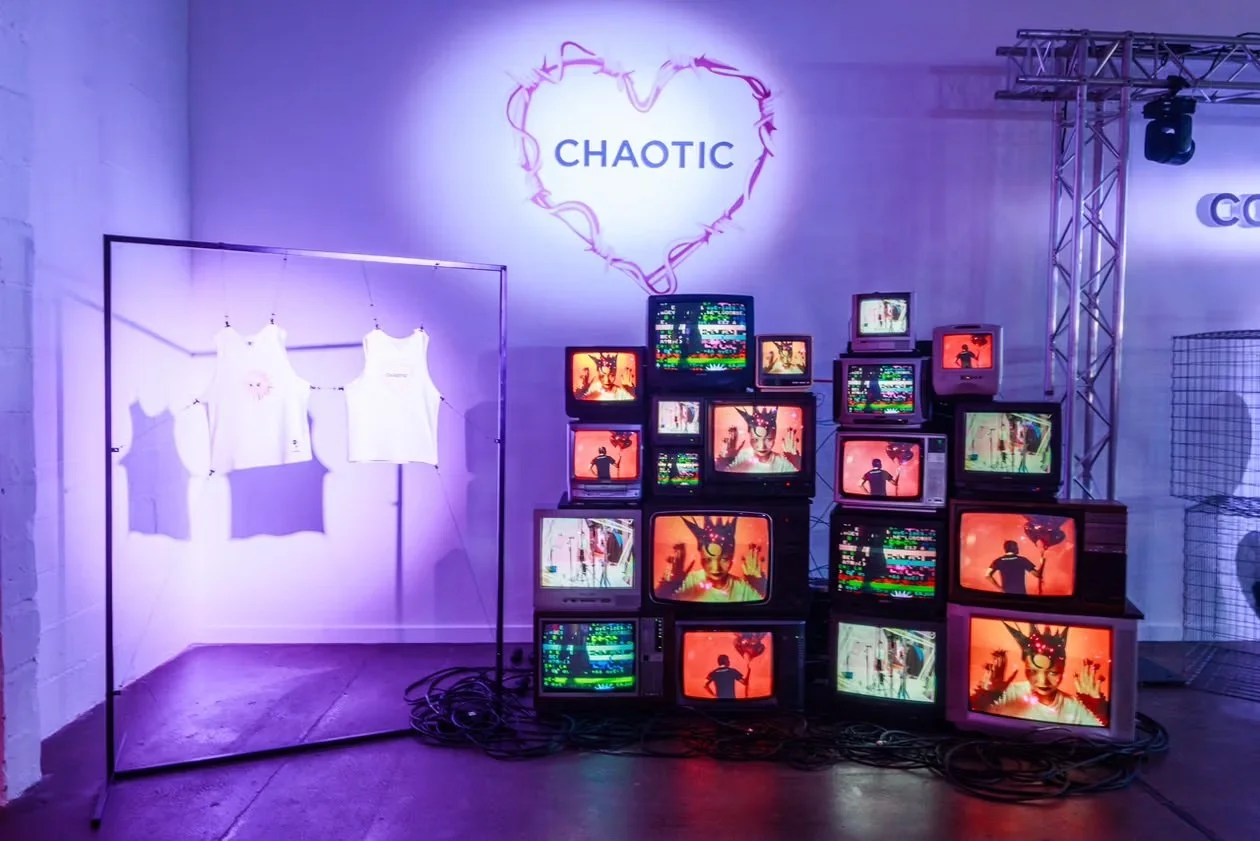 An art installation titled "Chaotic" features a wall with the word "Chaotic" inside a heart-shaped neon outline, a clothing display of white tank tops on a rack, and a collection of vintage CRT televisions stacked on the floor showing colorful digita