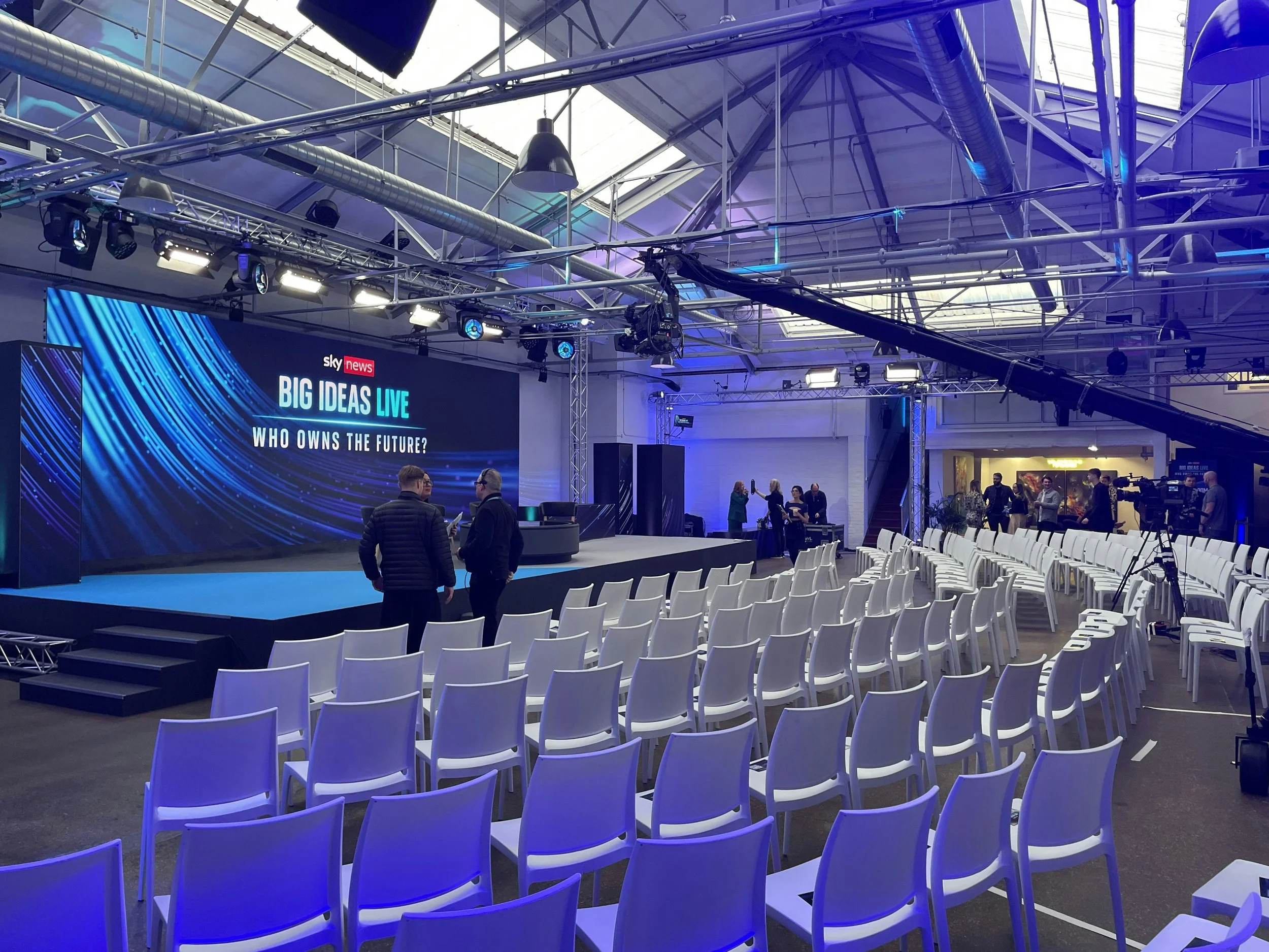 An event space set up for a TV broadcast or conference with rows of white chairs facing a stage with a large screen that reads 'Sky News Big Ideas Live: Who owns the future?'. There are a few people on and near the stage, and camera equipment is visi