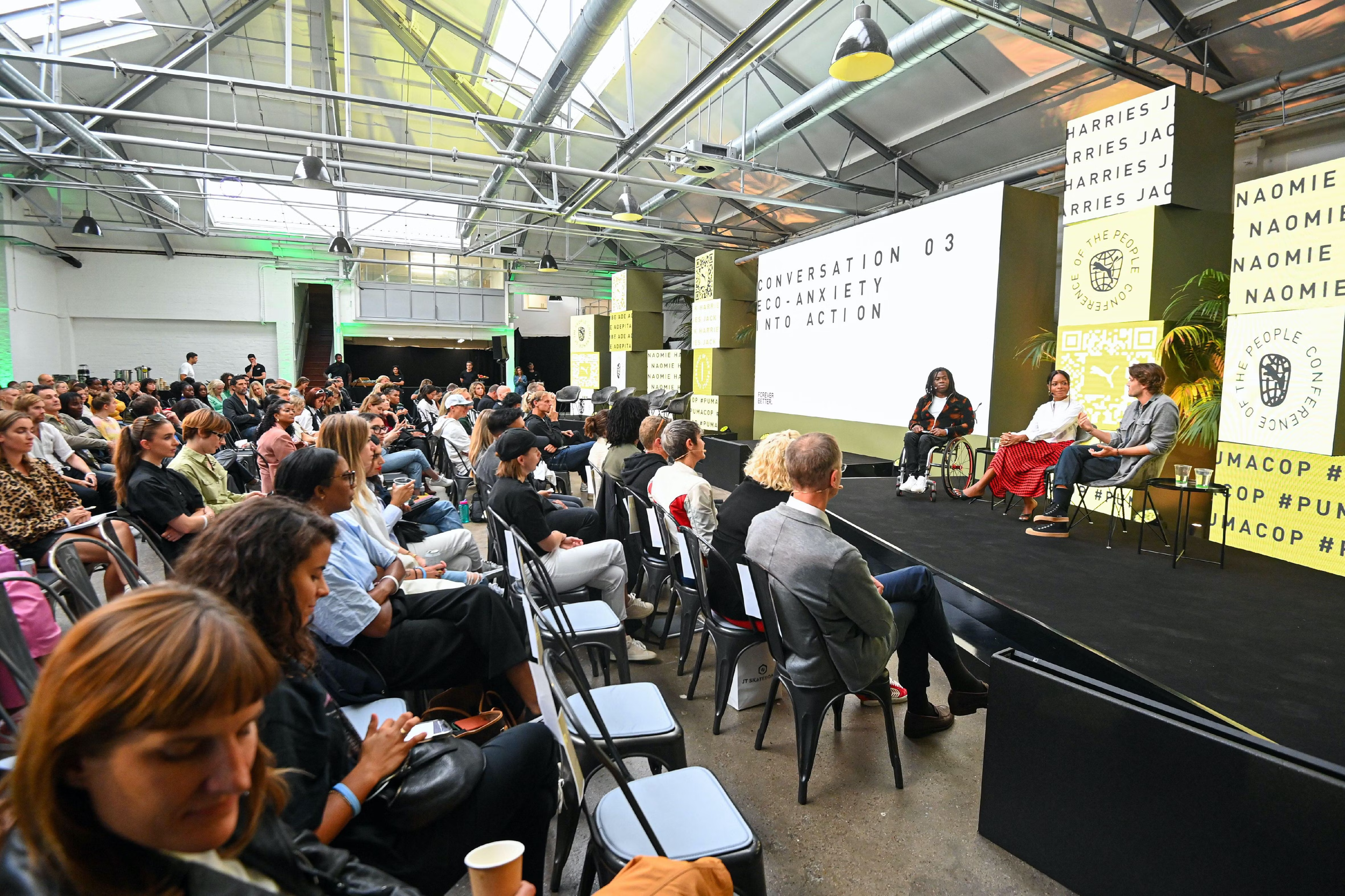 A large audience seated in front of a stage at a conference or panel discussion. Two speakers, one in a wheelchair, are seated on stage with a large screen behind them displaying the event theme, 'Conversation 03, Eco-Anxiety Into Action.' The stage 