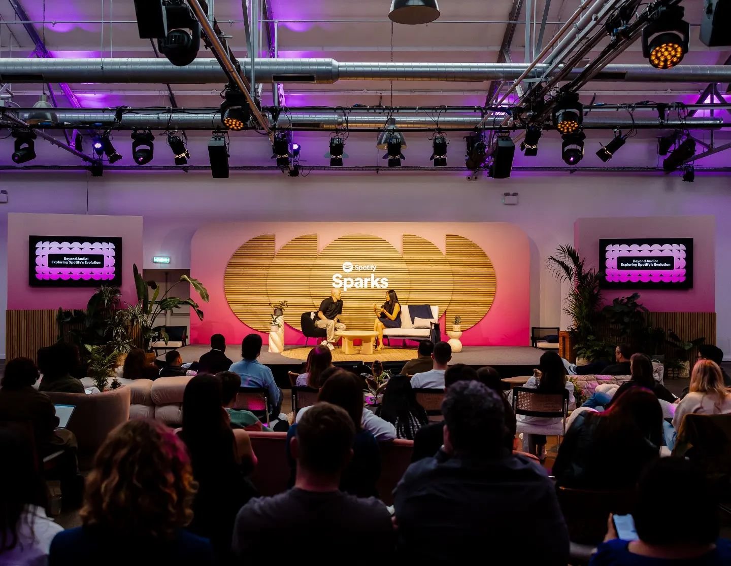 A stage with two people seated in conversation during a panel discussion, featuring a backdrop with the Spotify Sparks logo and two screens displaying the event title, with an audience watching in a modern venue with purple and pink lighting.