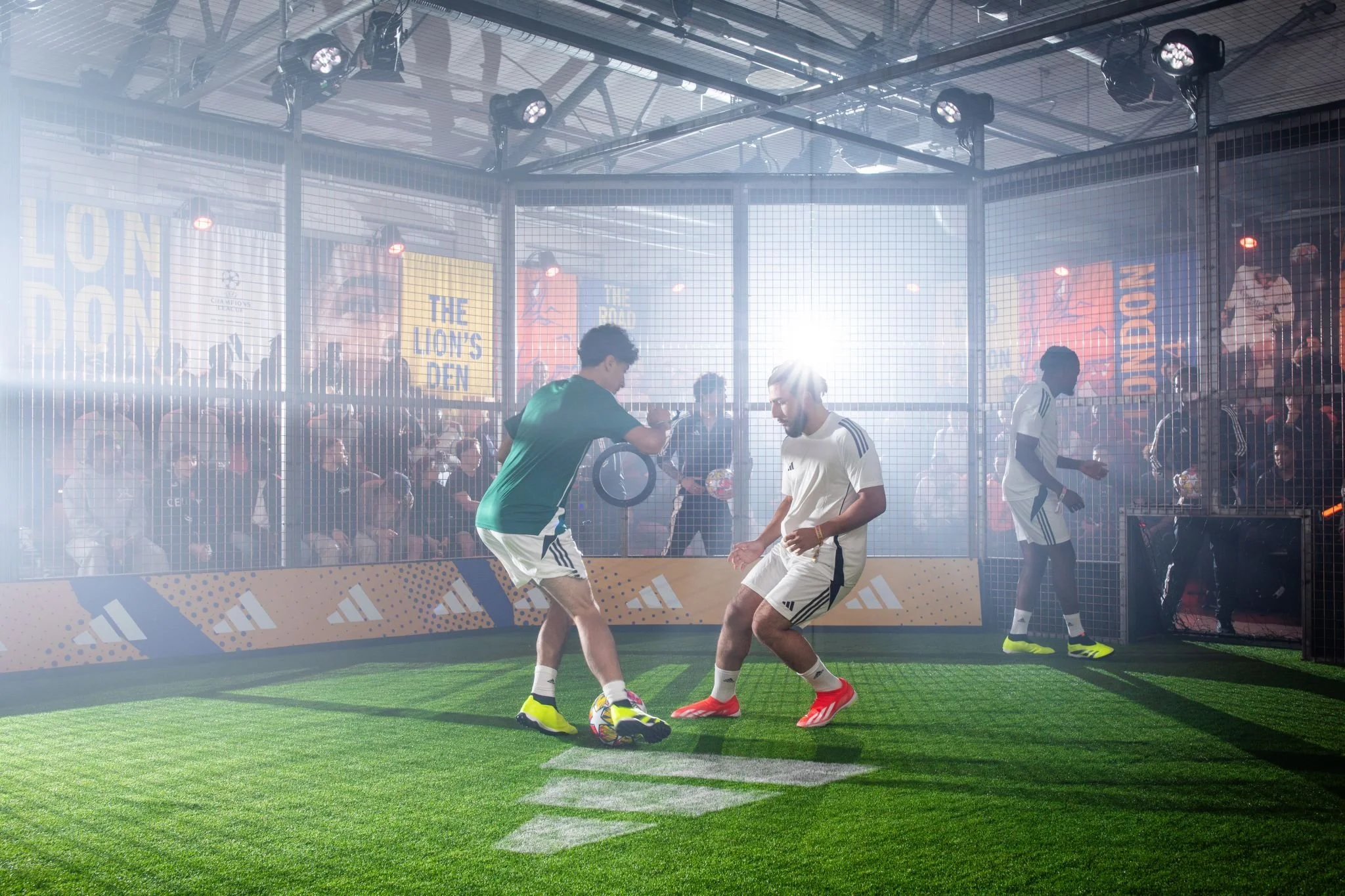 Two soccer players are practicing inside a small enclosed arena with artificial grass. They are in front of a crowd of spectators seated behind a netted barrier, with sunlight shining through the back.