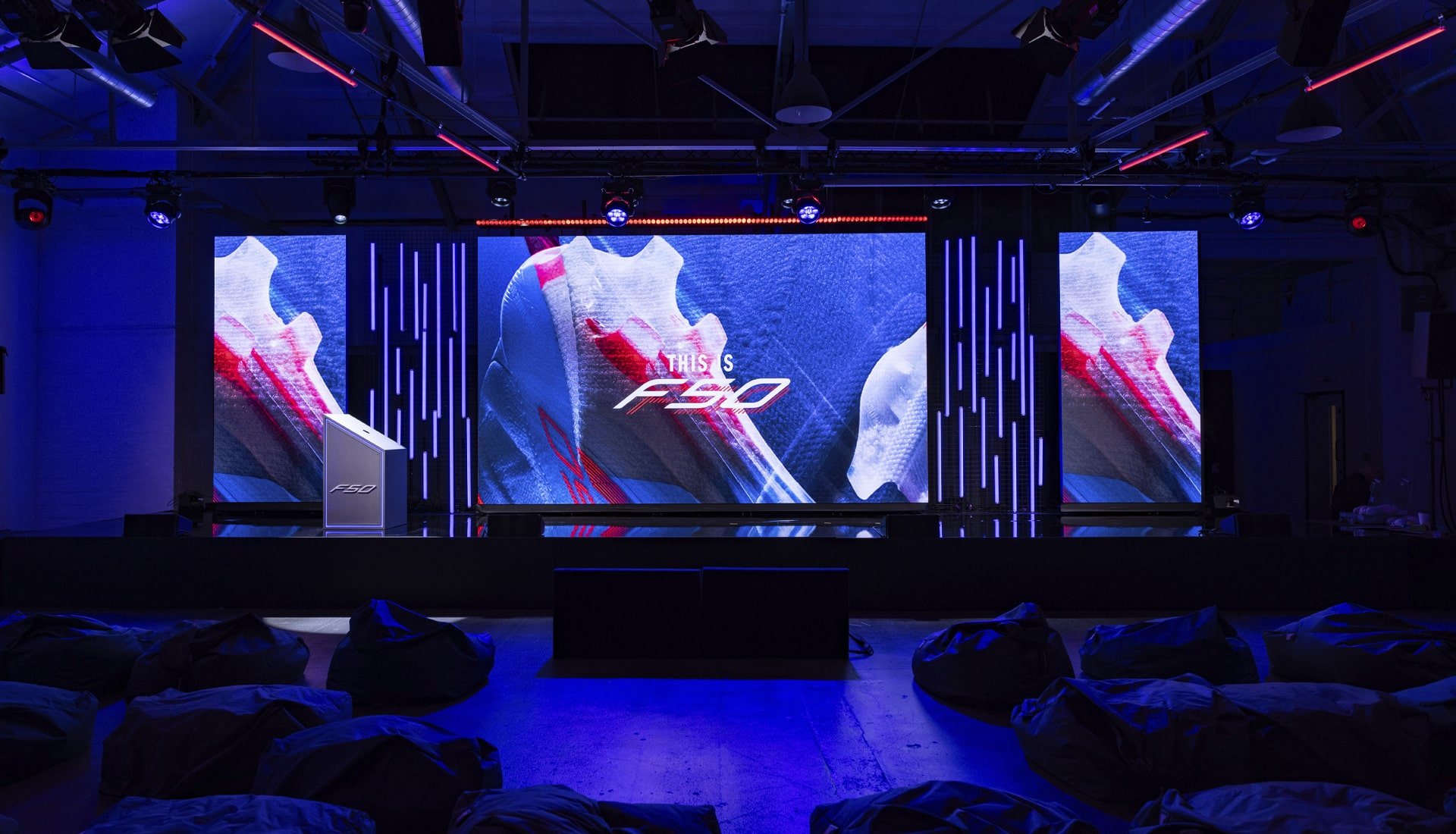 Stage set up for an event with large LED screens displaying a close-up image of a shoe and the logo 'F50'. The stage has blue and red lighting, bean bags on the floor, and a podium on the left.