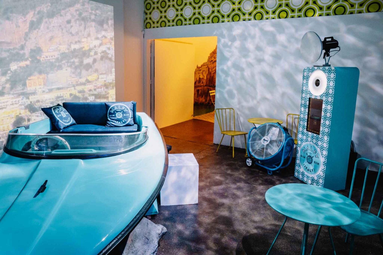 Interior of a room with a vintage boat, yellow chairs, a fan, and a gin vending machine with Shelfy Girl branding, illuminated by colorful and decorative walls.