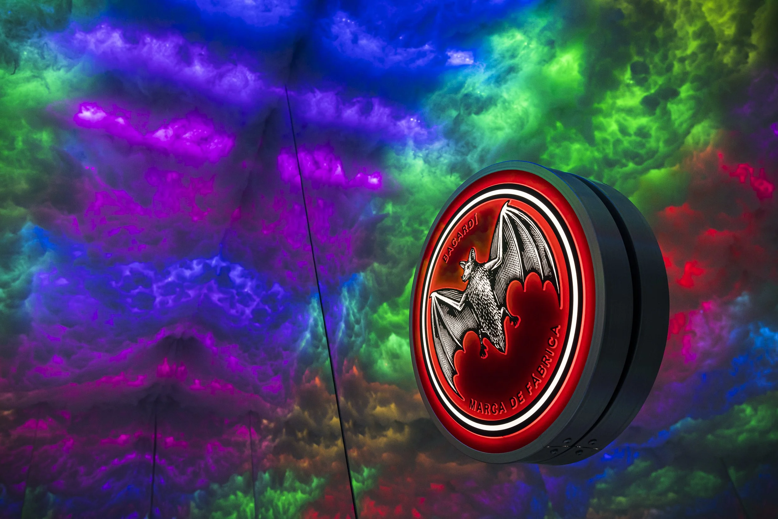 A round red and black illuminated sign with a bat logo and the text "BAGARDI MARCA DE FABRICA" is shown against a colorful, swirling cloudy sky background.