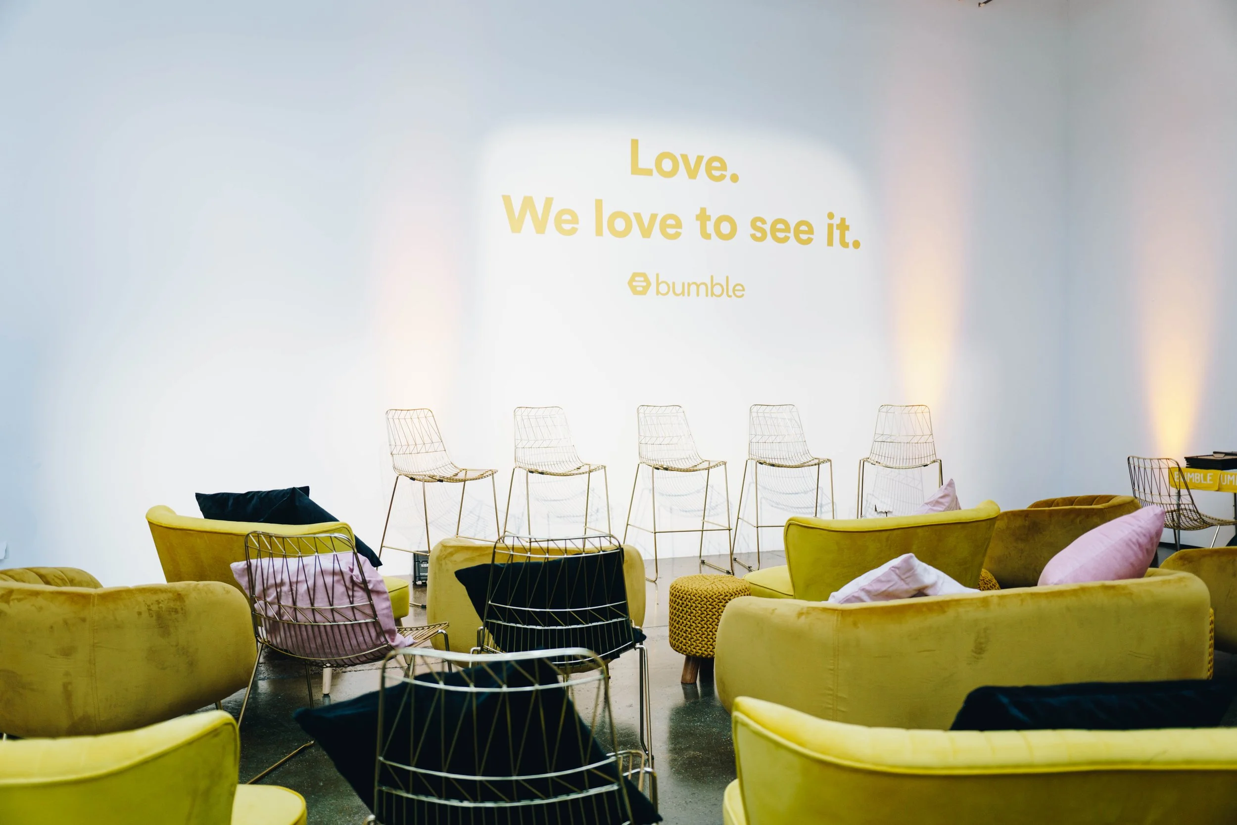 A brightly lit studio space in Protein Studios, with yellow sofas and pink pillows, arranged facing a white wall with yellow text that reads "Love. We love to see it." and the Bumble logo.