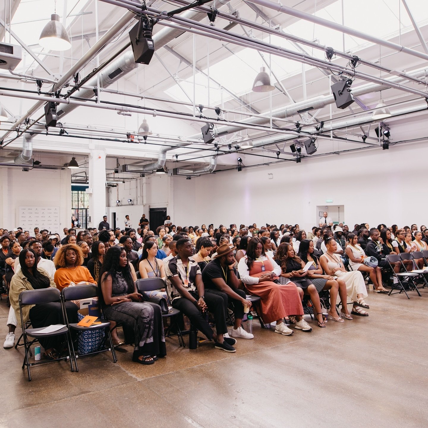 A large group of people seated in a spacious, white, industrial-style venue with high ceilings, led lighting, and exposed ducts, attending an event or presentation.