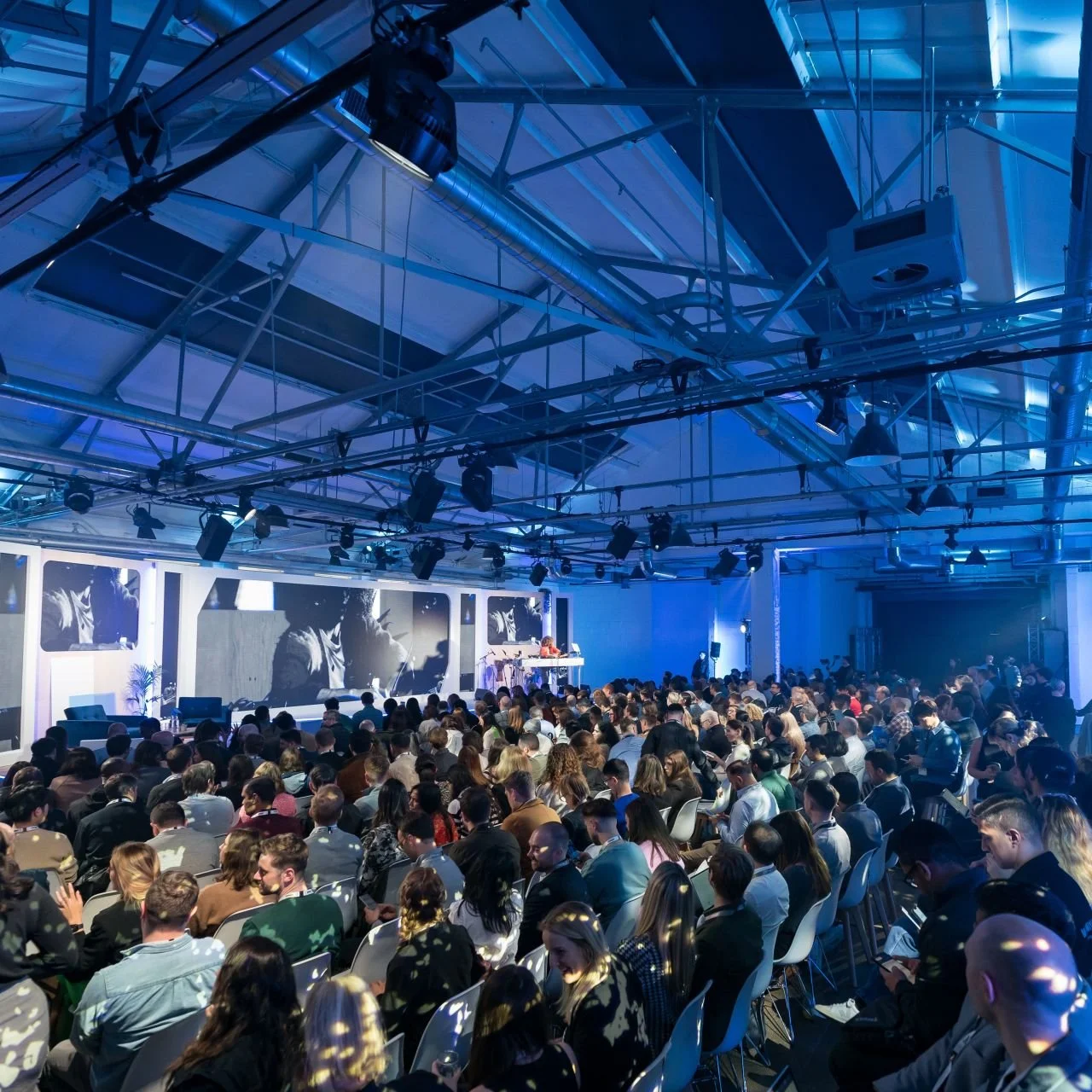 Protein Studios hosting a large indoor conference with a stage, large screens, and a seated audience facing the stage, illuminated by blue lighting.