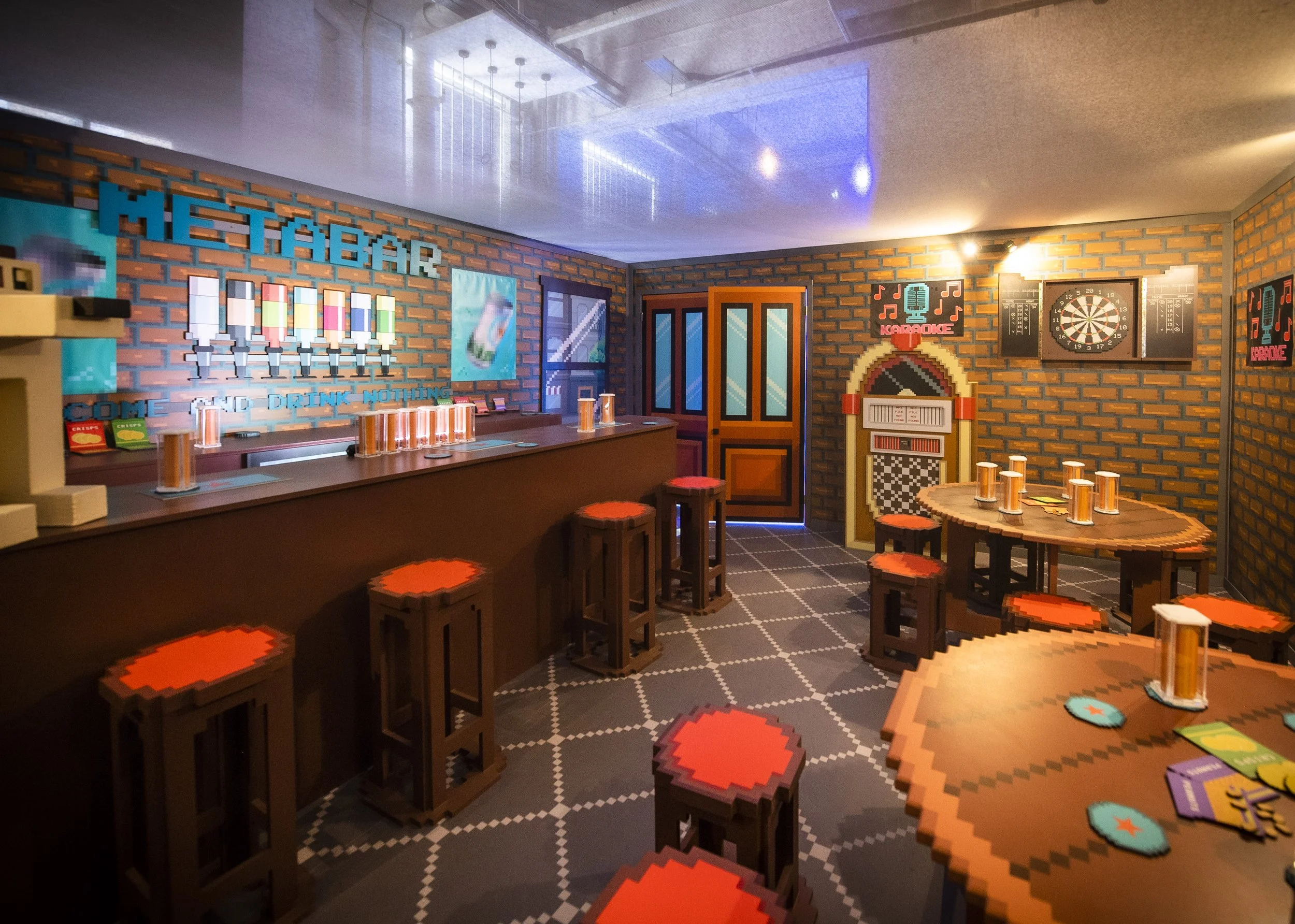 A lively bar and game room with a brick interior, featuring a dartboard, karaoke sign, and round tables with stools. The bar has tall stools, colorful posters, and a patterned floor.