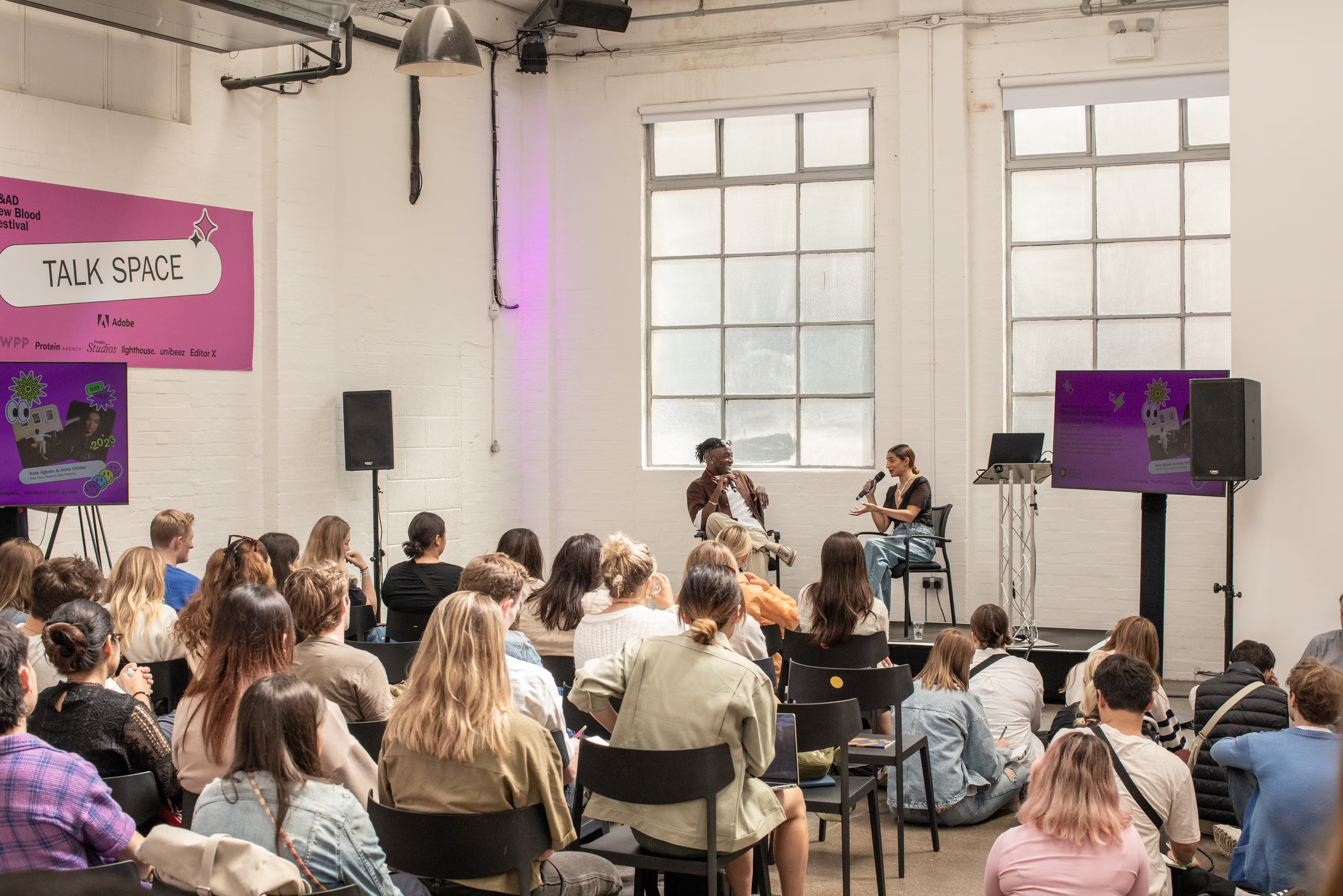 A group of people attending a talk or discussion in a bright room with large windows and high ceilings. Two individuals are seated on a small stage, one holding a microphone, engaging in conversation while the audience listens.