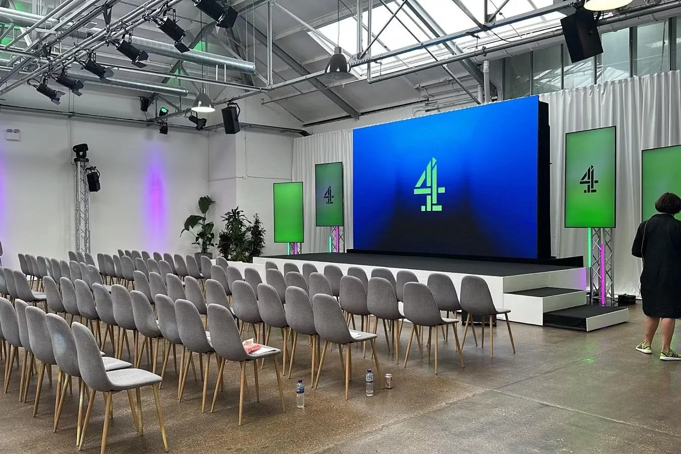Empty conference room with rows of gray chairs facing a large blue screen with the number 4 on it, three smaller screens with the same logo on green backgrounds, and stage lighting in an industrial-style space with plants and a woman standing on the 