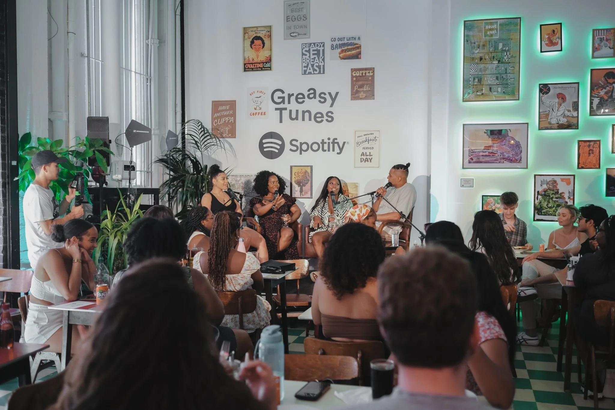 A diverse group of people gathered in a cafe or event space with a mural on the wall that reads 'Greasy Tunes' and 'Spotify.' A panel of four women and one man are seated on stage, speaking into microphones. The audience is engaged, with some taking 