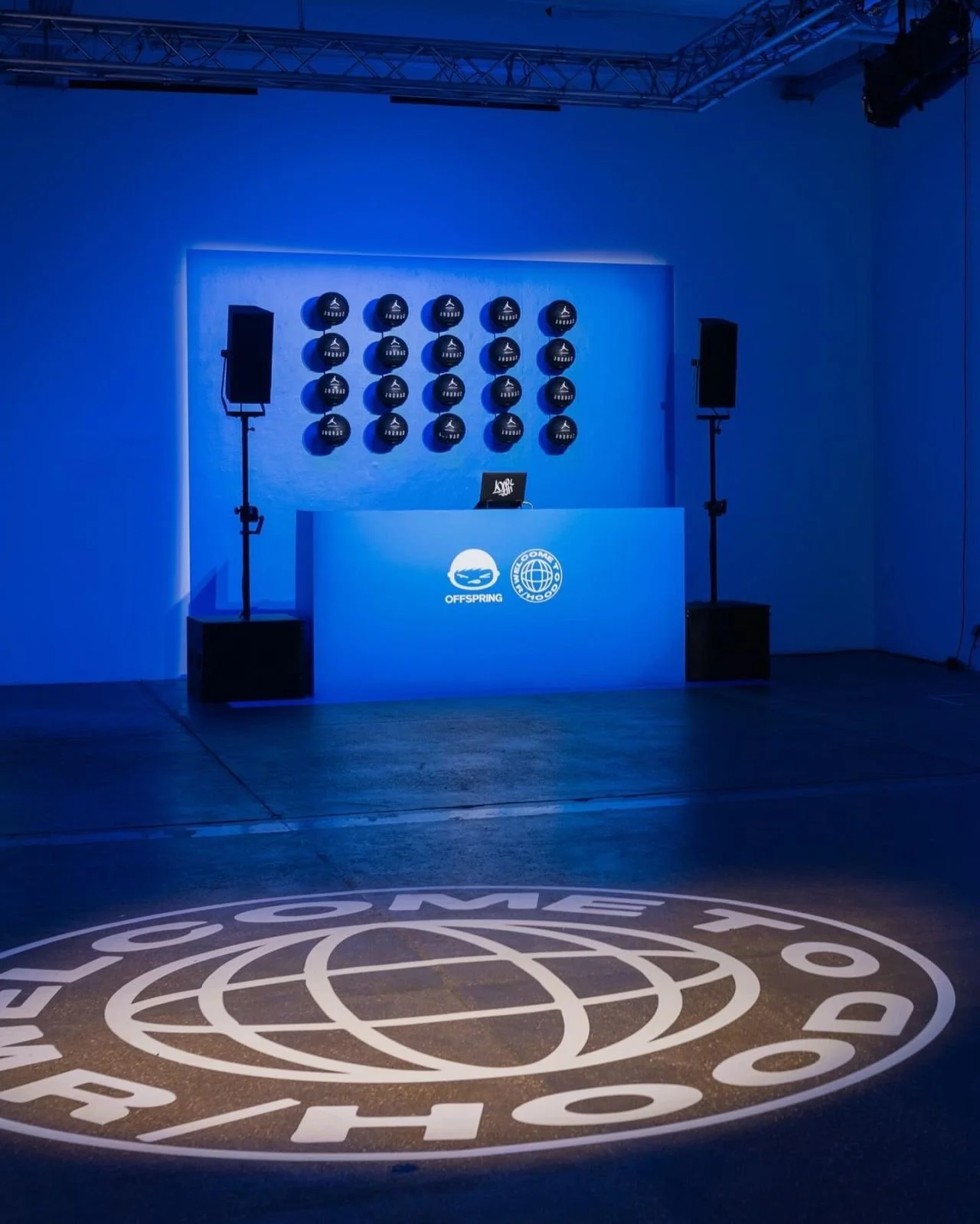 An indoor stage setup with speakers and laptop, illuminated in blue lighting, featuring a large circular logo on the floor that reads 'OFFSPRING' with a globe and a stylized triangle.