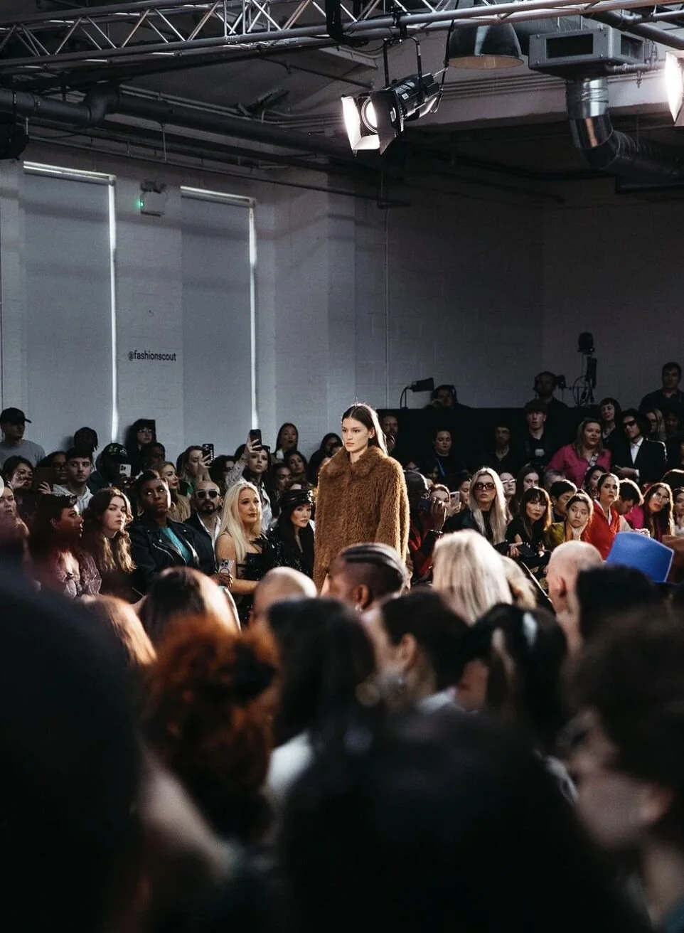 Fashion model walking down the runway during a fashion show, surrounded by an audience seated in front of her.