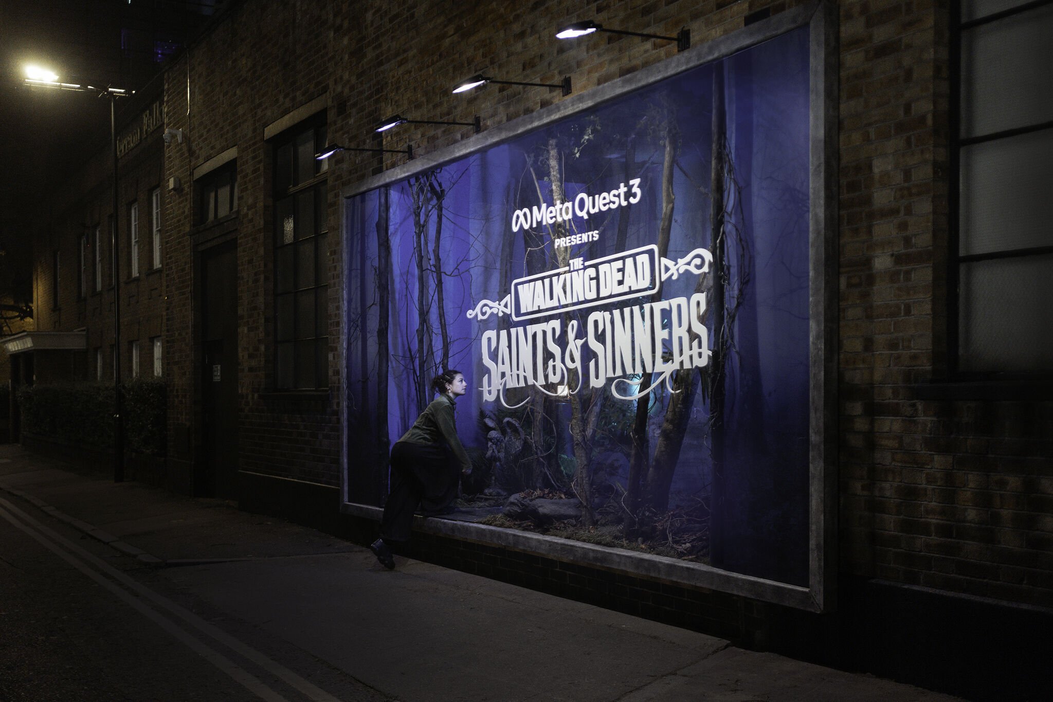 A person wearing a green jacket and black pants kneeling in front of a large outdoor advertisement for 'The Walking Dead: Saints & Sinners' game by Meta Quest 3, displayed on a window of a brick building at night with street lights.