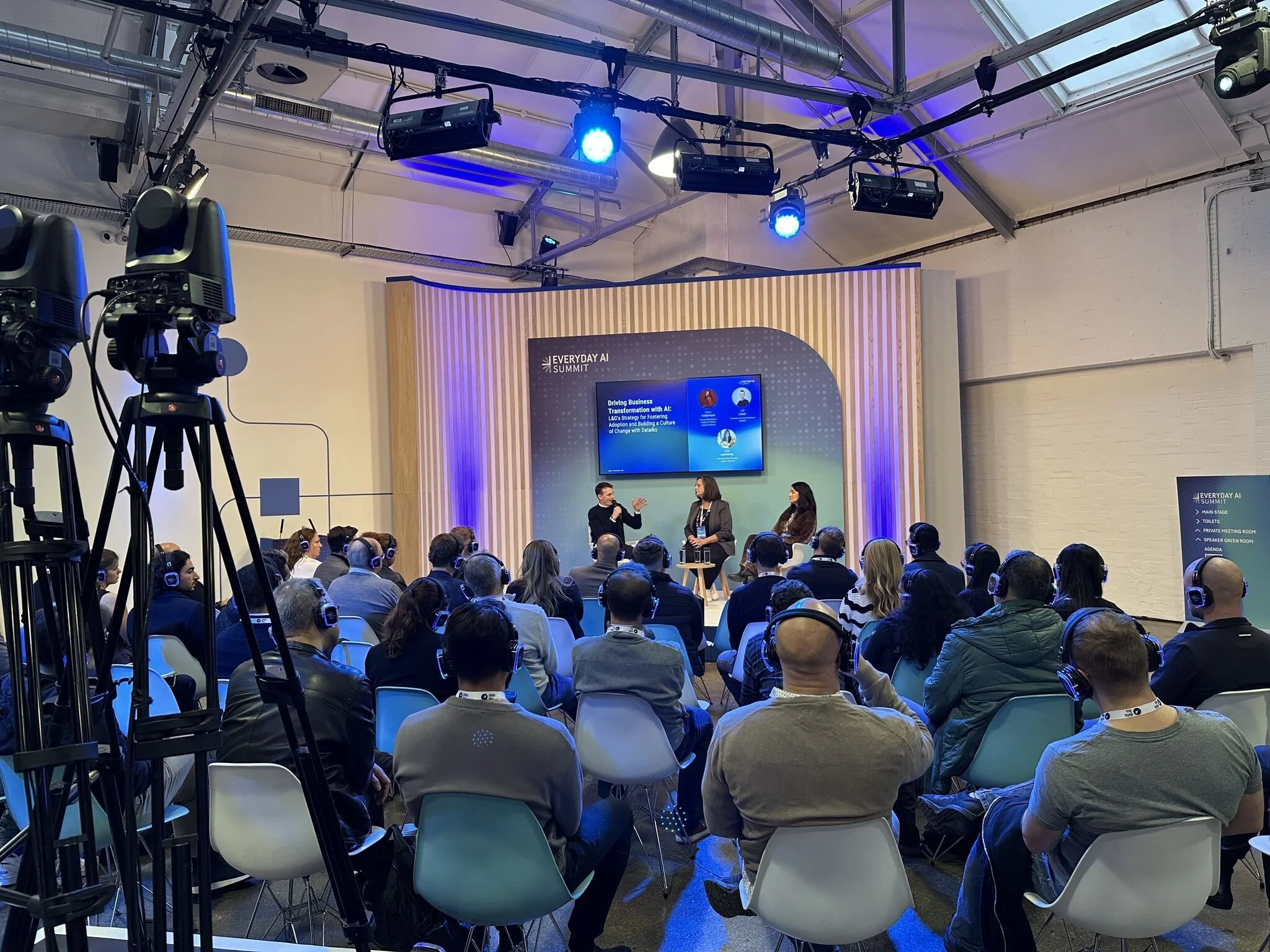 Audience attending a conference or seminar with headphones on, listening to speakers on stage at the Everyday AI Summit, with cameras recording the event.