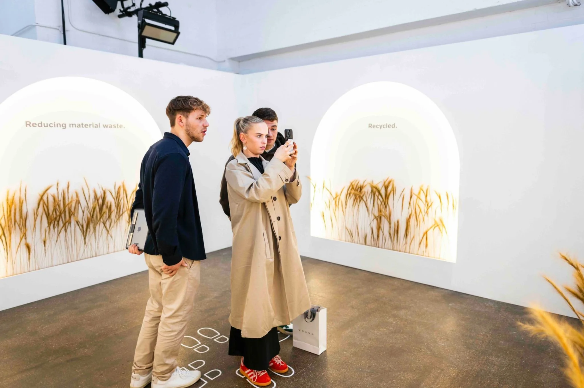 Three people standing in an art gallery, observing wheat stalks on display with text about recycling and reducing waste. One person is taking a photo with a smartphone.