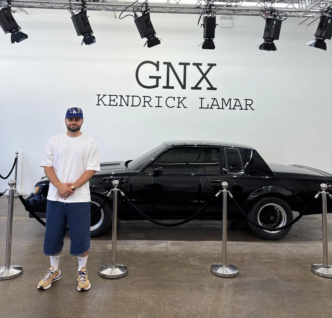 An attendee standing in front of a black luxury car at an exhibit with the text 'GNX Kendrick Lamar' displayed on the wall behind him.