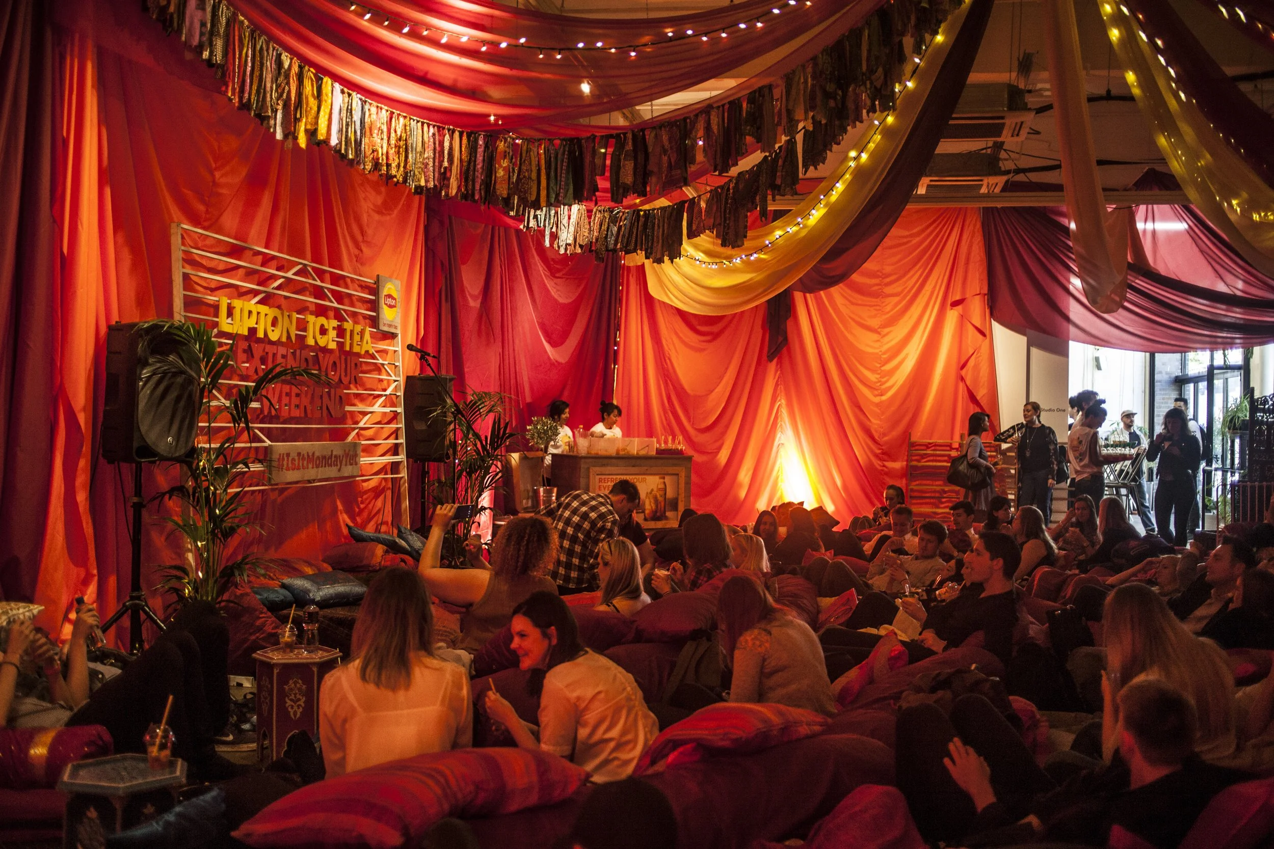 A lively indoor event with warm red, orange, and yellow drapery and string lights. A stage with two women, a DJ or host, and a small crowd seated on cushions and chairs, socializing and enjoying the atmosphere.