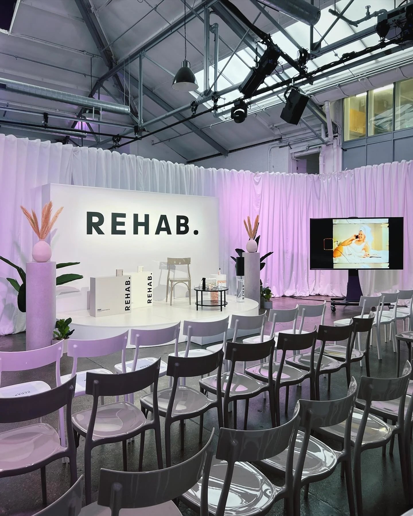 Event space with a stage, chairs, and a large sign that says 'REHAB.' with decorative plants. There is a TV screen showing a person in bed, and white curtains around the area.