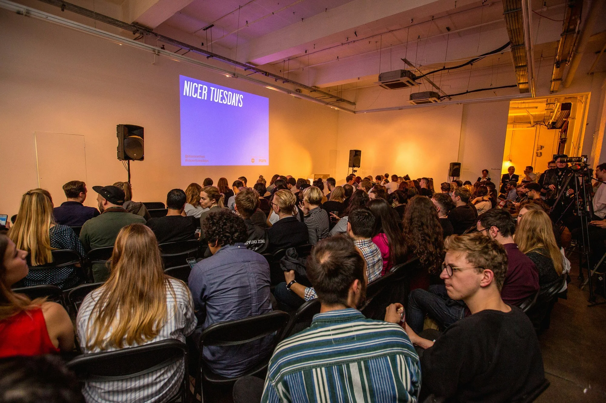 Audience seated in a large conference room with a presentation slide on the wall that reads 'Nicer Tuesdays'.