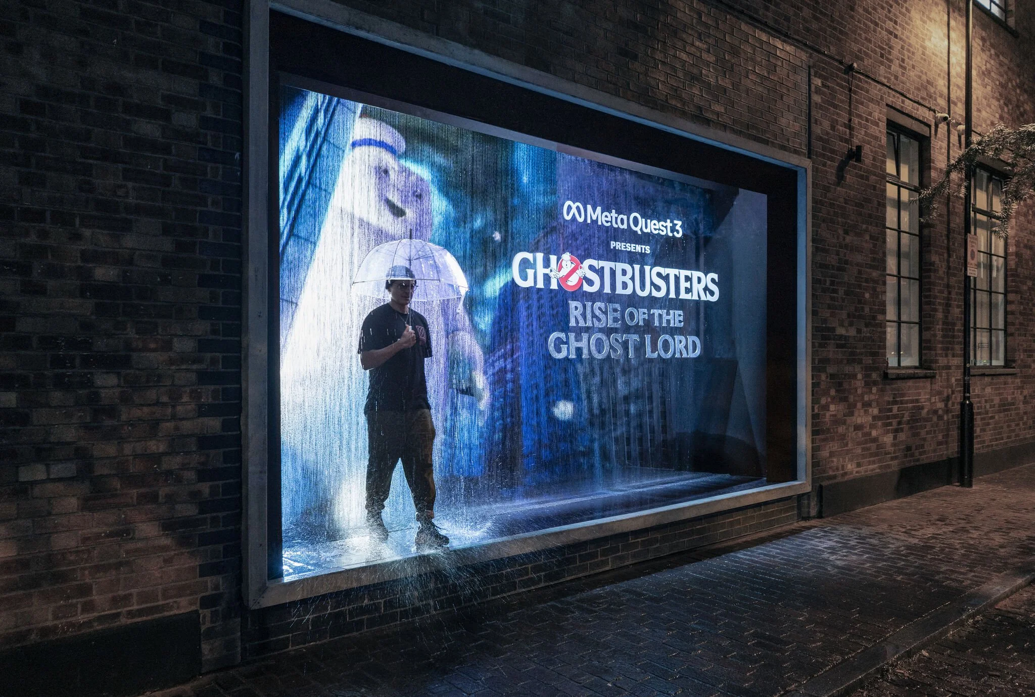 A person holding a transparent umbrella inside a display window for the movie Ghostbusters: Rise of the Ghost Lord, sponsored by Meta Quest 3, with rain falling on the window and a cartoon ghost in the background.