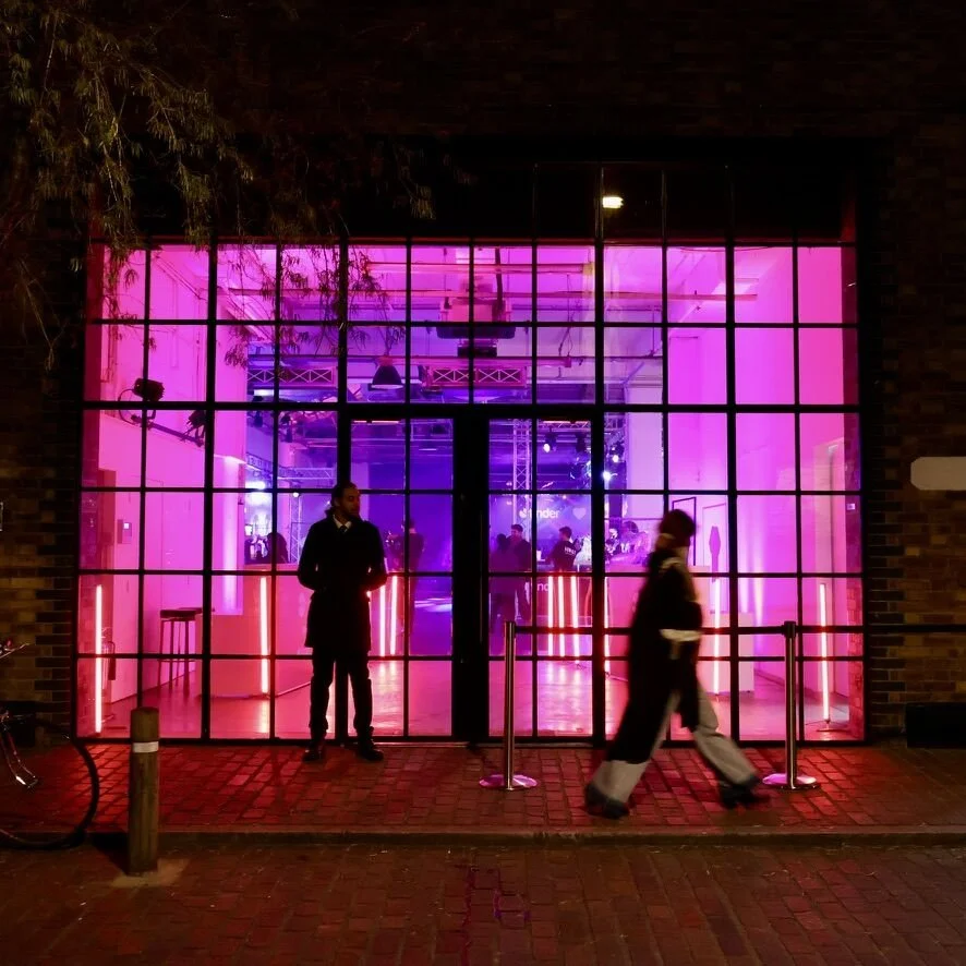 Night view of a building with large glass windows illuminated with pink and purple neon lights, showing an interior with people and stage lighting.