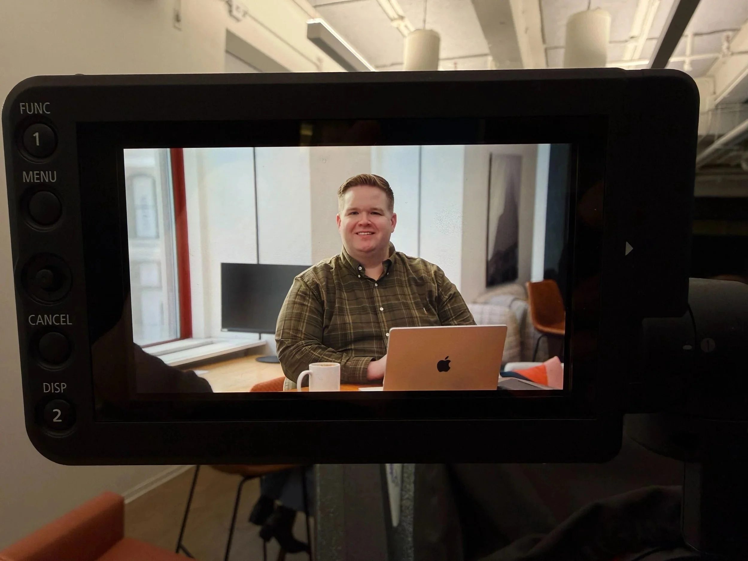 Andrew Seaman, a man sitting at a table with a laptop in front of him, smiling and wearing a plaid shirt, photographed through a camera screen.