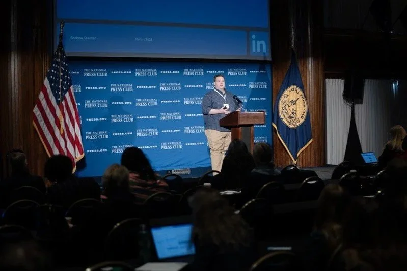 Andrew Seaman giving a speech at a podium at the National Press Club, with American flag and a blue Kentucky state flag behind them, and an audience seated in front.
