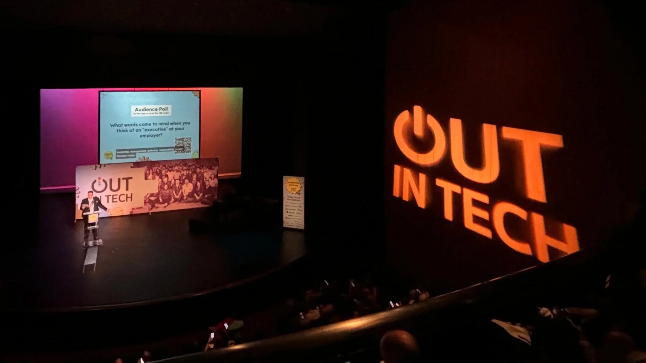 Andrew Seaman speaking at a tech conference with a large screen displaying an audience poll and a speaker standing at a podium. The background has a logo that reads 'OUT IN TECH' in large illuminated letters.