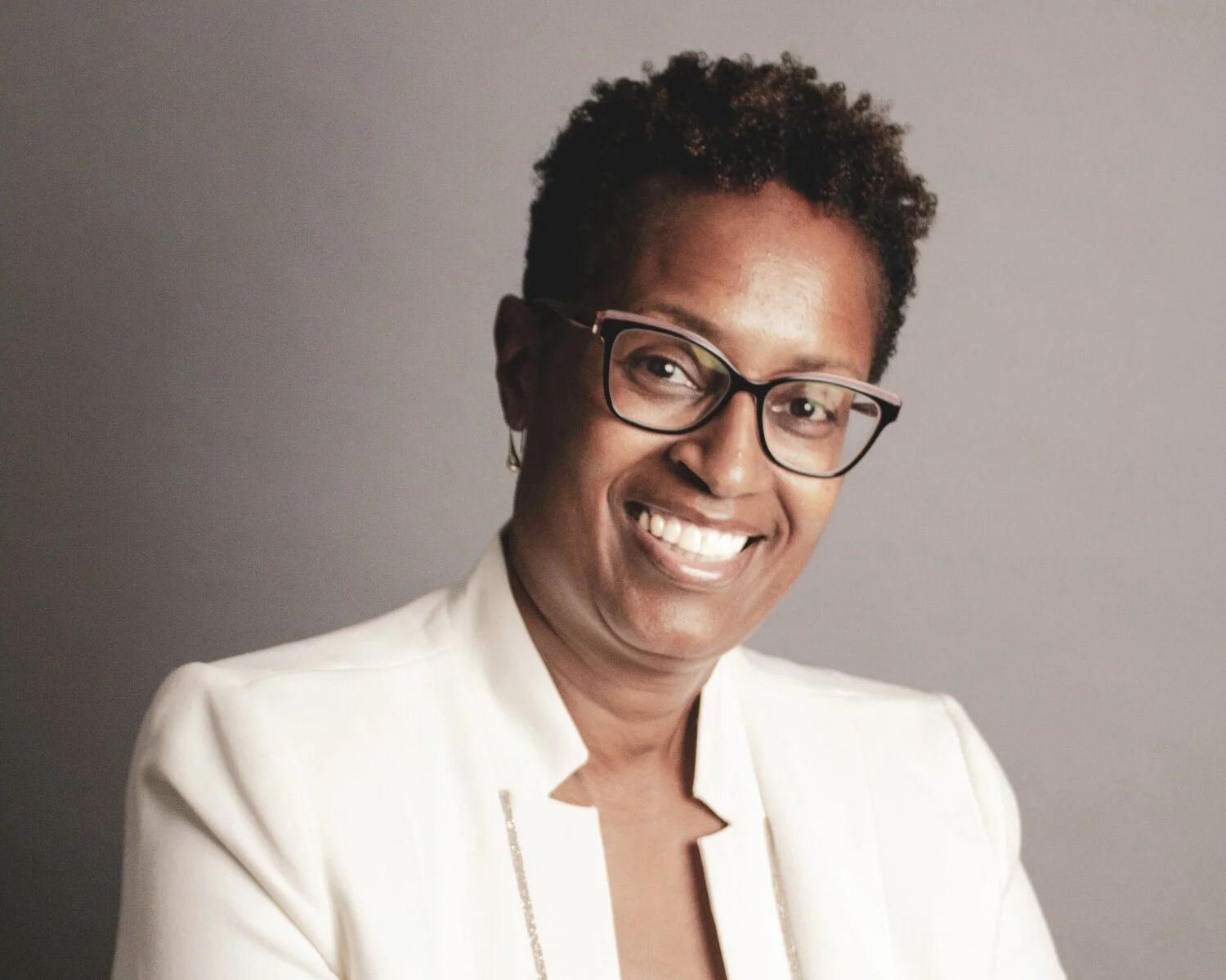 Person wearing glasses and a white jacket smiling at the camera with a neutral background.