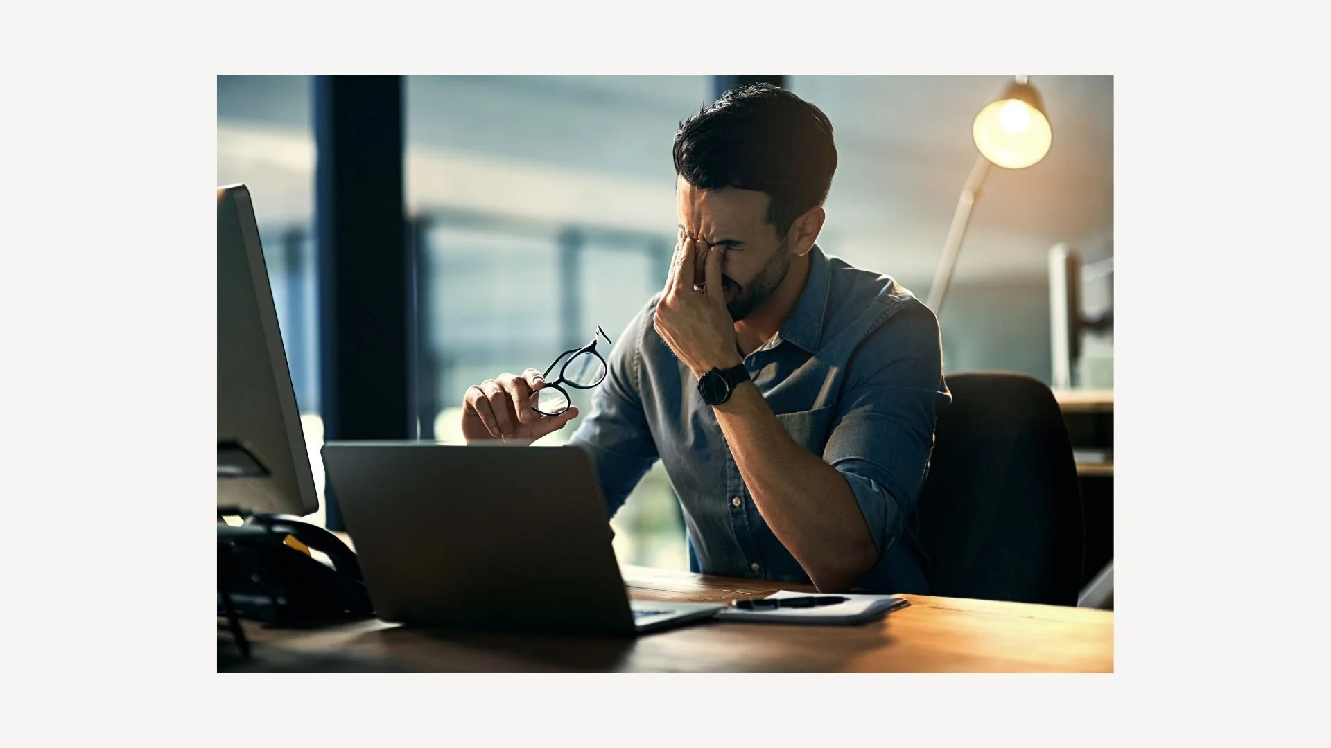 Man sitting at desk with laptop, holding glasses, looking stressed, rubbing eyes.