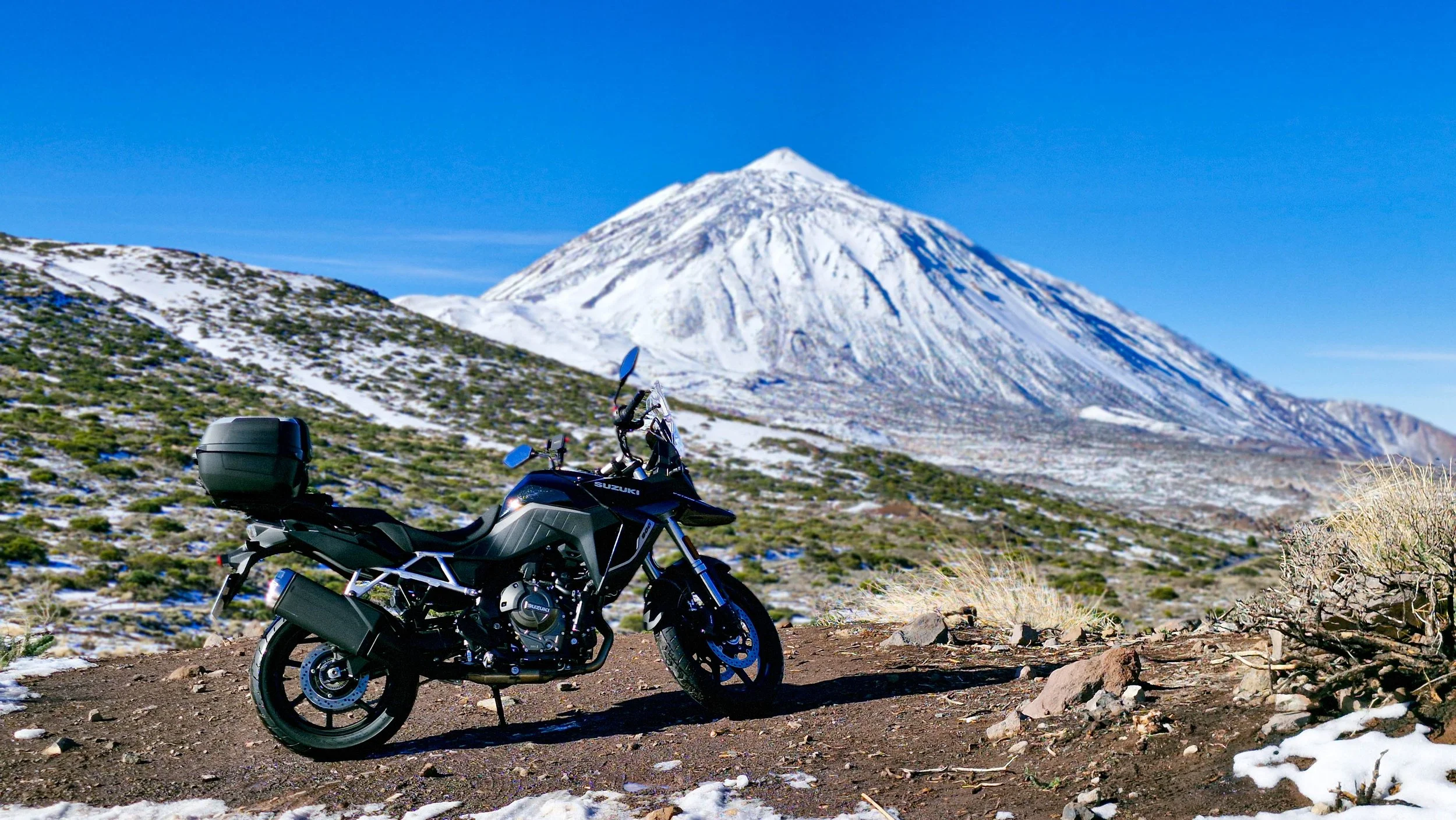 Una motocicletta nera e grigia parcheggiata in un paesaggio montuoso innevato, con un vulcano che si erge sullo sfondo.