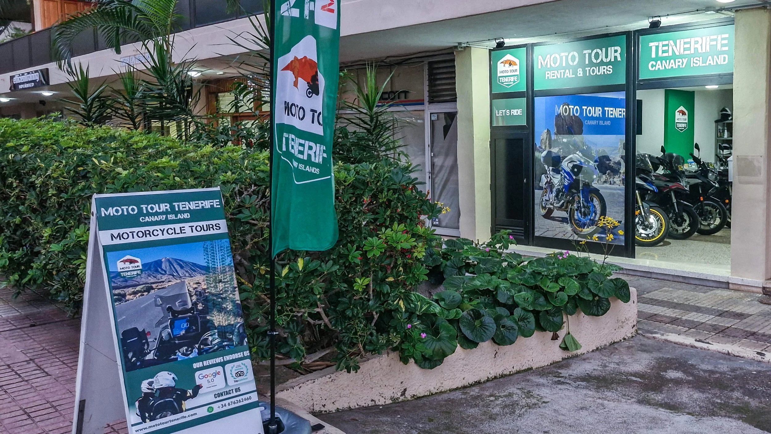Fachada de tienda de alquiler y tours en moto en Tenerife, Canarias, con motocicletas en exhibición y señalización promocional para tours por la isla. Rodeada de plantas y arbustos en la acera.