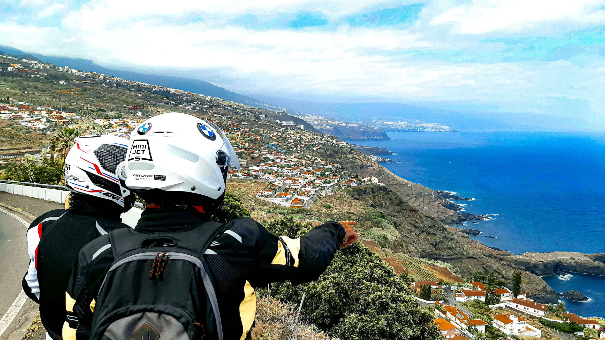 Dos motociclistas con cascos y mochilas observando la vista de un paisaje costero con casas, vegetación y el océano.