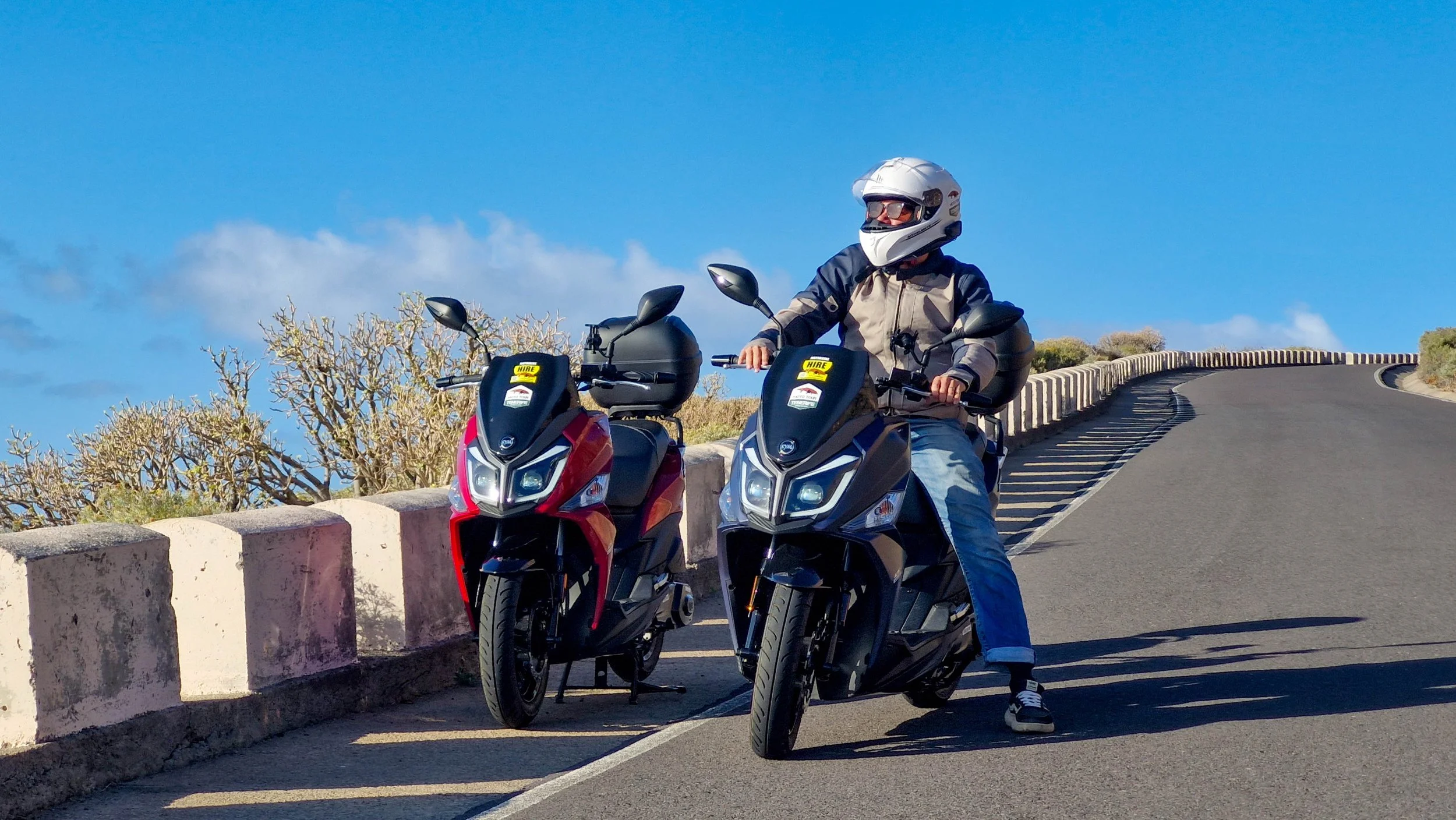 Due persone con casco e giubbotto protettivo in sella a uno scooter su una strada con una barriera e della vegetazione sullo sfondo.