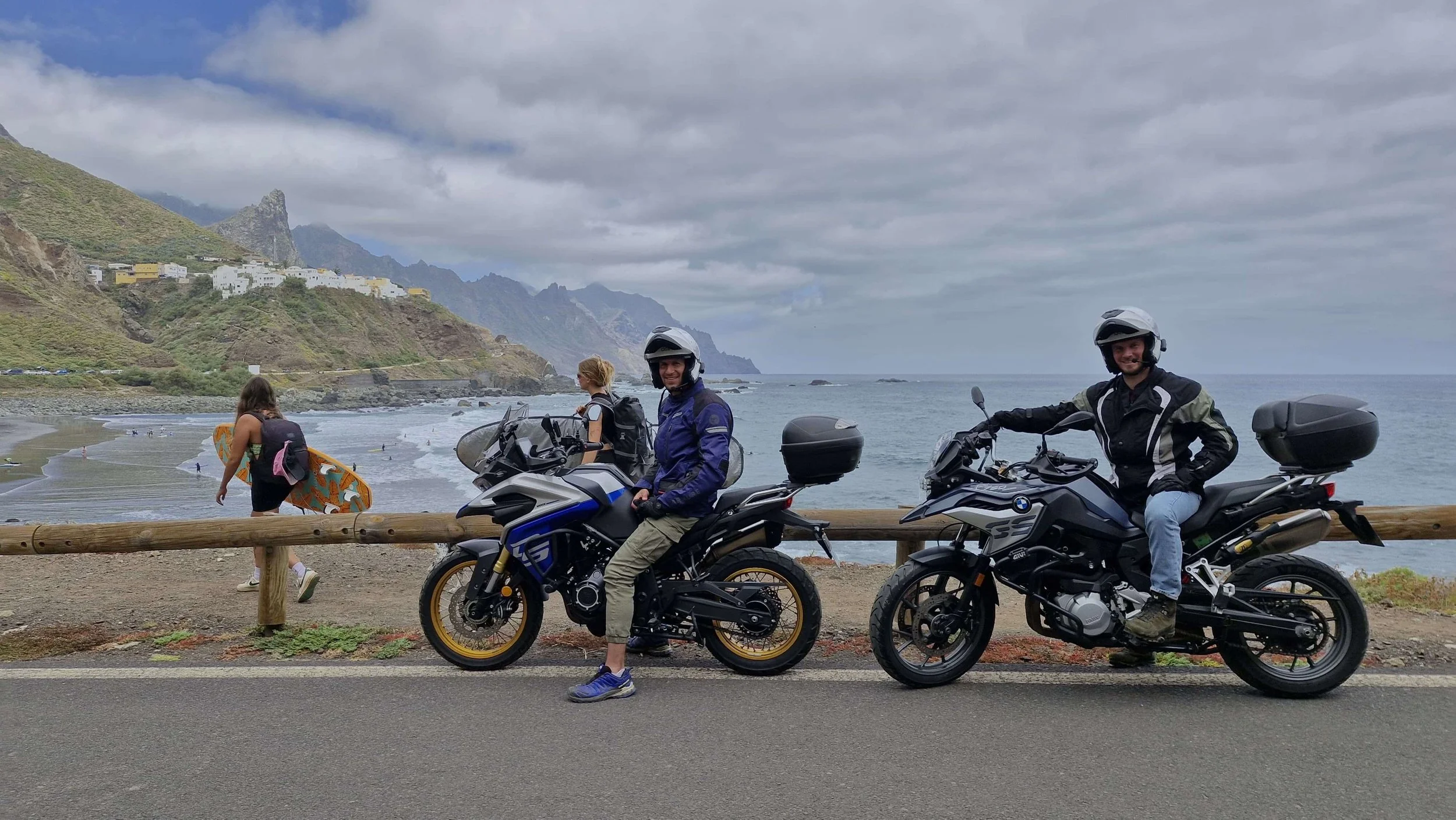 Dos motociclistas con cascos descansando junto a la costa con mar y montañas, una persona con gorra caminando con patineta en la playa.
