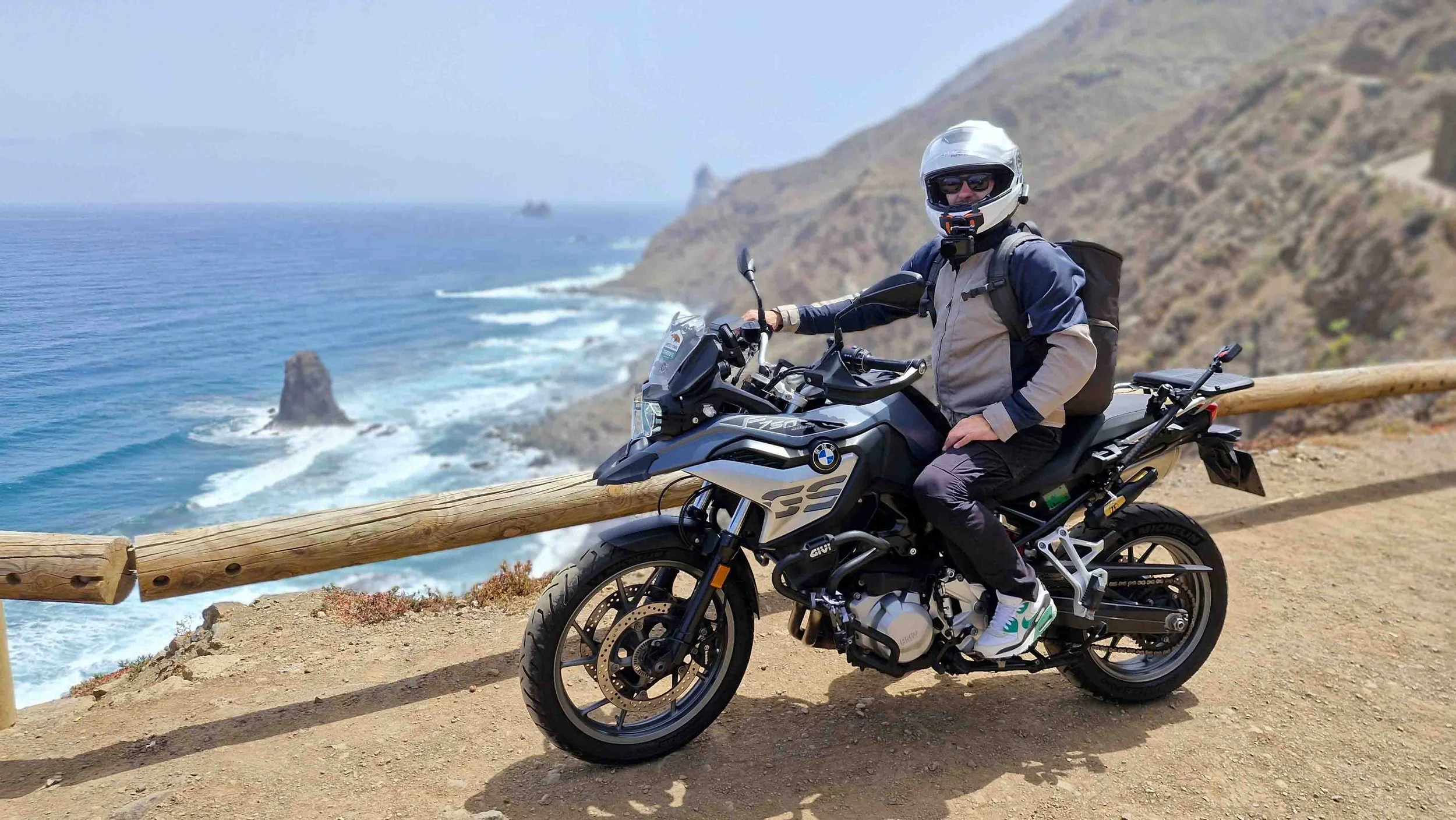 Una persona con casco, occhiali e abbigliamento da motociclista seduta su una moto BMW, accanto a una barriera di legno su una strada costiera con scogliere e una formazione rocciosa nell'acqua.