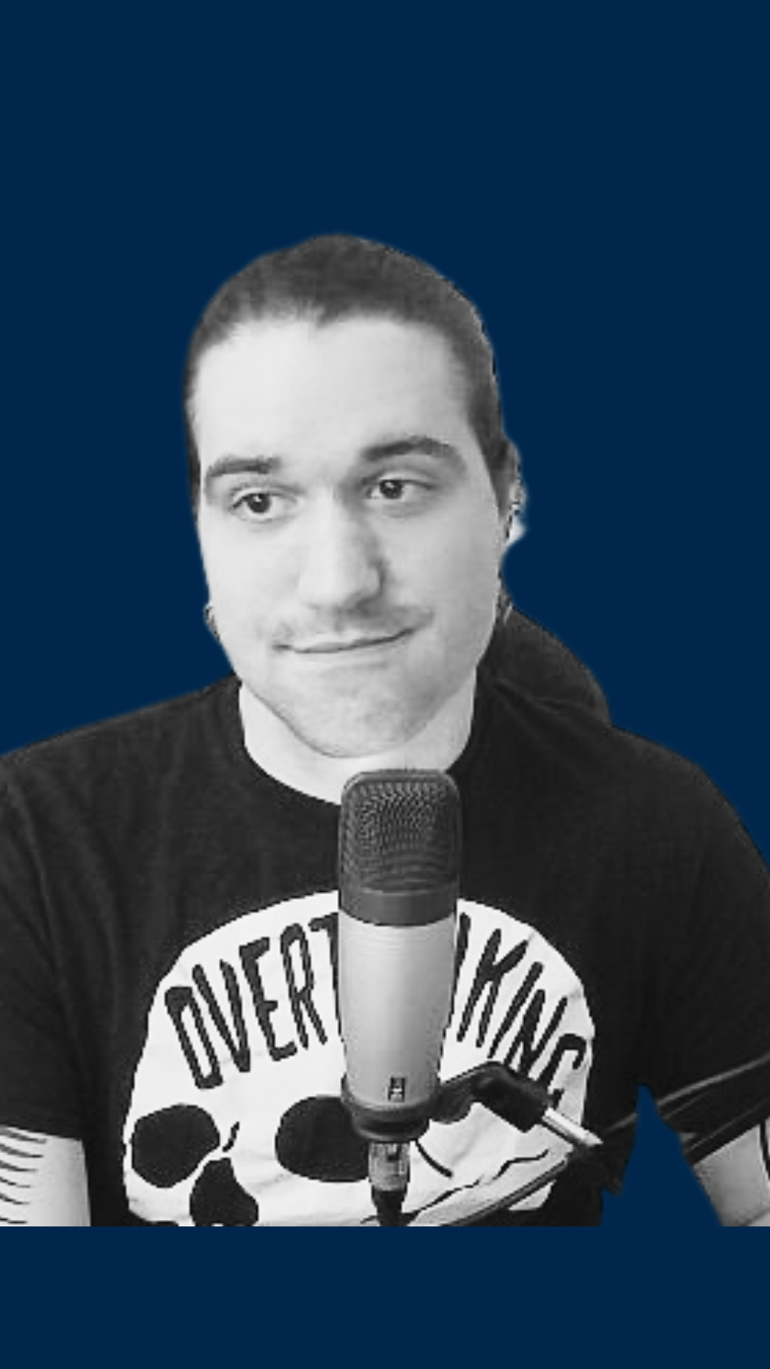 Black-and-white photo of a man with short hair, wearing a black T-shirt with a skull graphic and text, speaking into a microphone against a monochrome blue background.