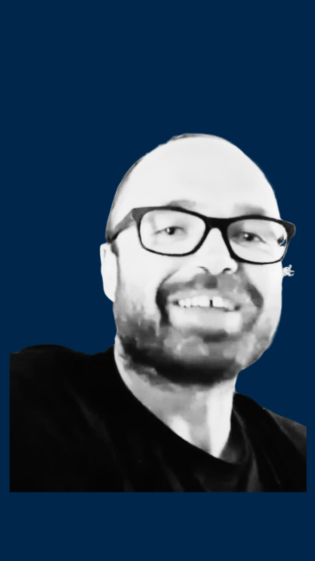 Black-and-white photo of a smiling man with glasses, a beard, and short hair, wearing a dark shirt, against a dark blue background.