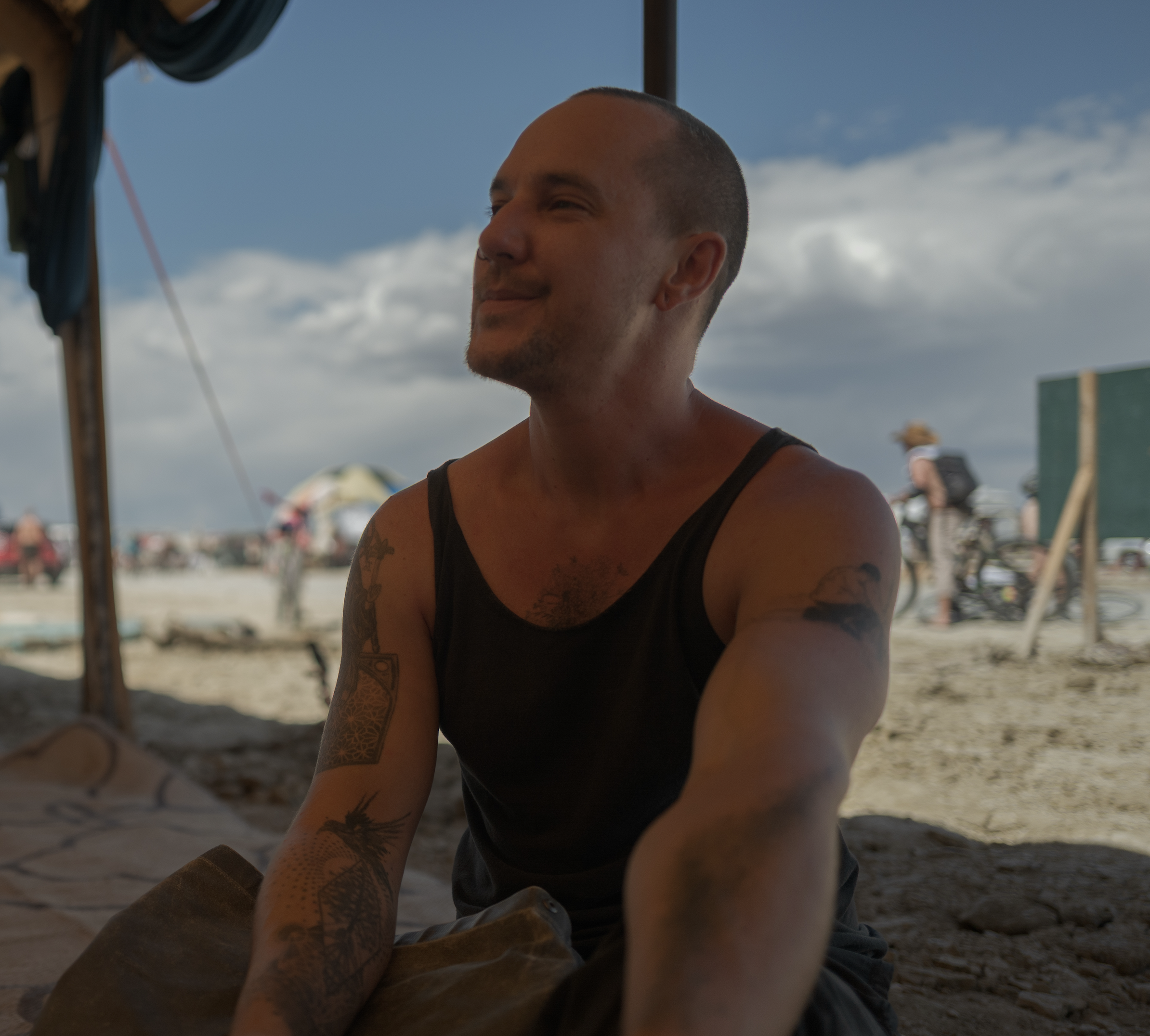 A man with tattoos sitting under a beach tent on a beach, smiling with eyes closed, in front of a partly cloudy sky.