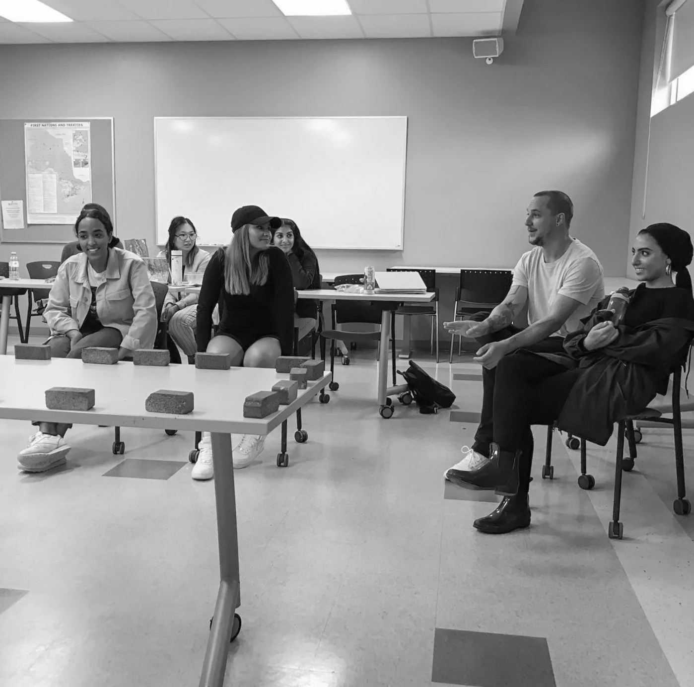 Group of young people sitting in a classroom, engaged in a discussion with a man who is speaking and gesturing with his hands. The classroom has projects on the desk, a whiteboard, and a map on the wall.