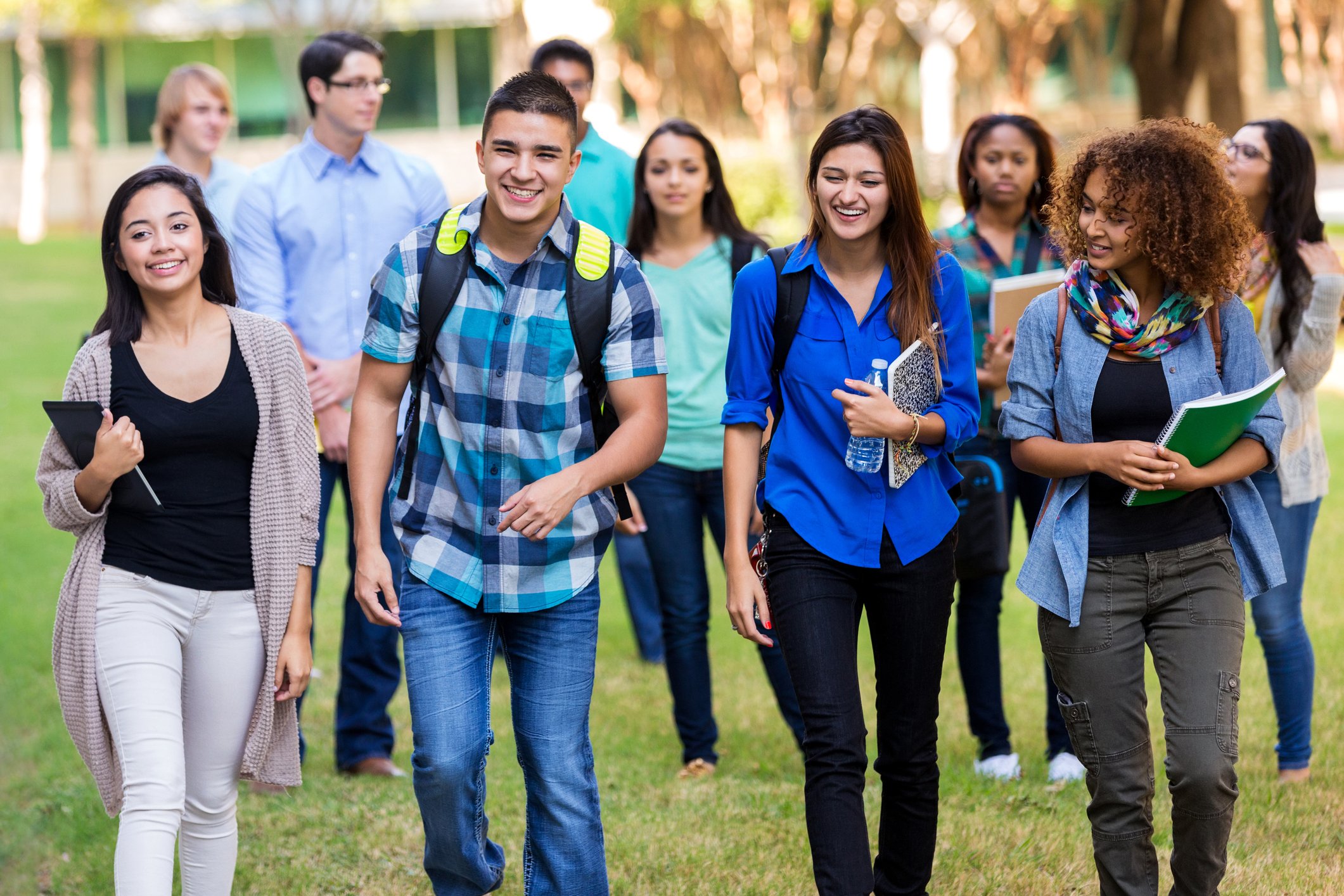 Group of diverse young students walking outside on a college campus, smiling and talking, carrying notebooks and water bottles.