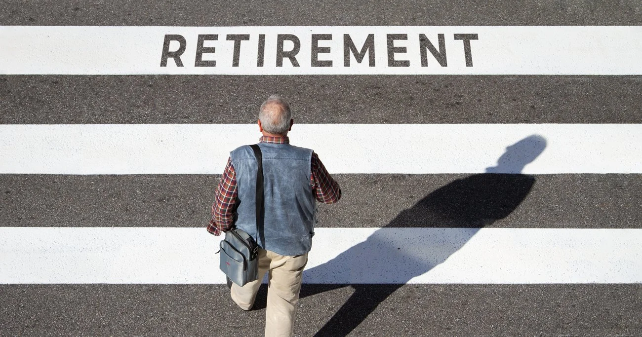 An older man walking across a crosswalk labeled with the word 'RETIREMENT'. His shadow is visible on the pavement.